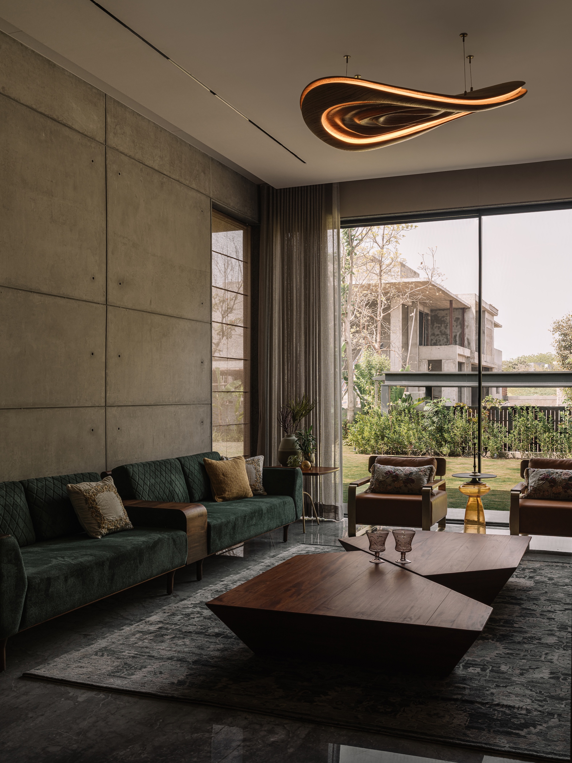 Living room with a green velvet sofa, faceted timber coffee table and glowing ceiling light beside a concrete panel wall and garden glazing