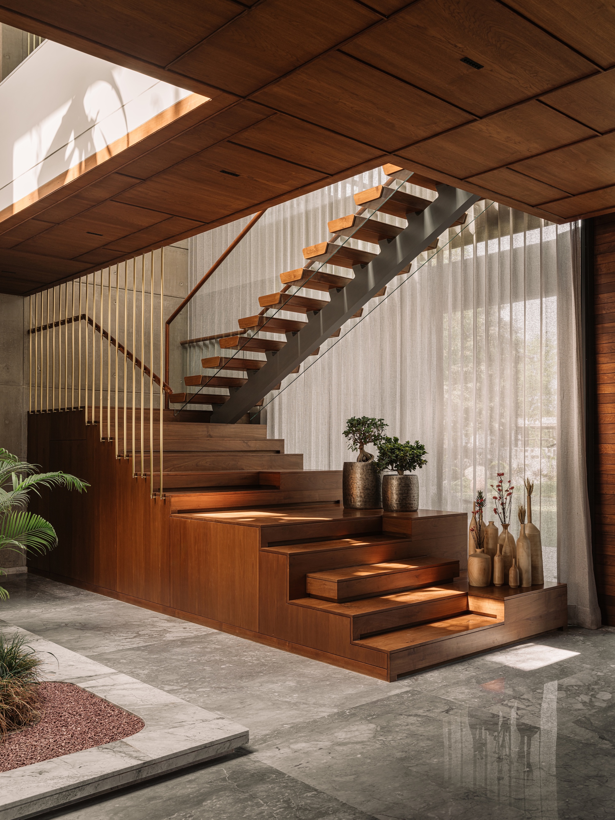 Timber staircase with floating treads and a steel stringer behind sheer curtains, afternoon sun striking the polished stone floor