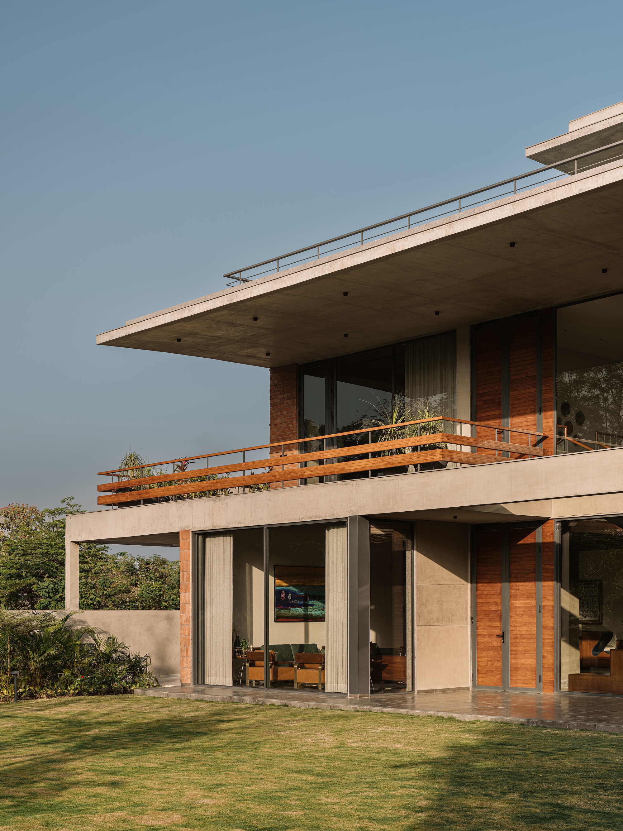 Side view of the glazed ground floor and timber-railed balcony beneath the concrete roof slab, with a sunlit lawn in front
