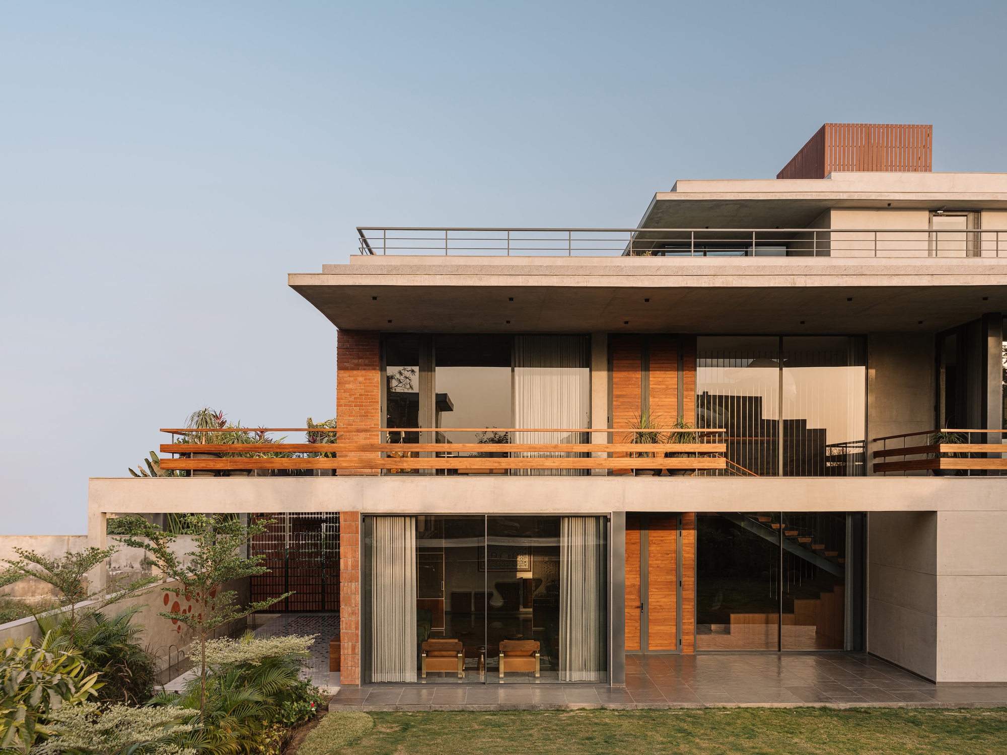 Garden facade with concrete slabs, brick piers and timber railings, glazed living room at ground level in soft evening haze