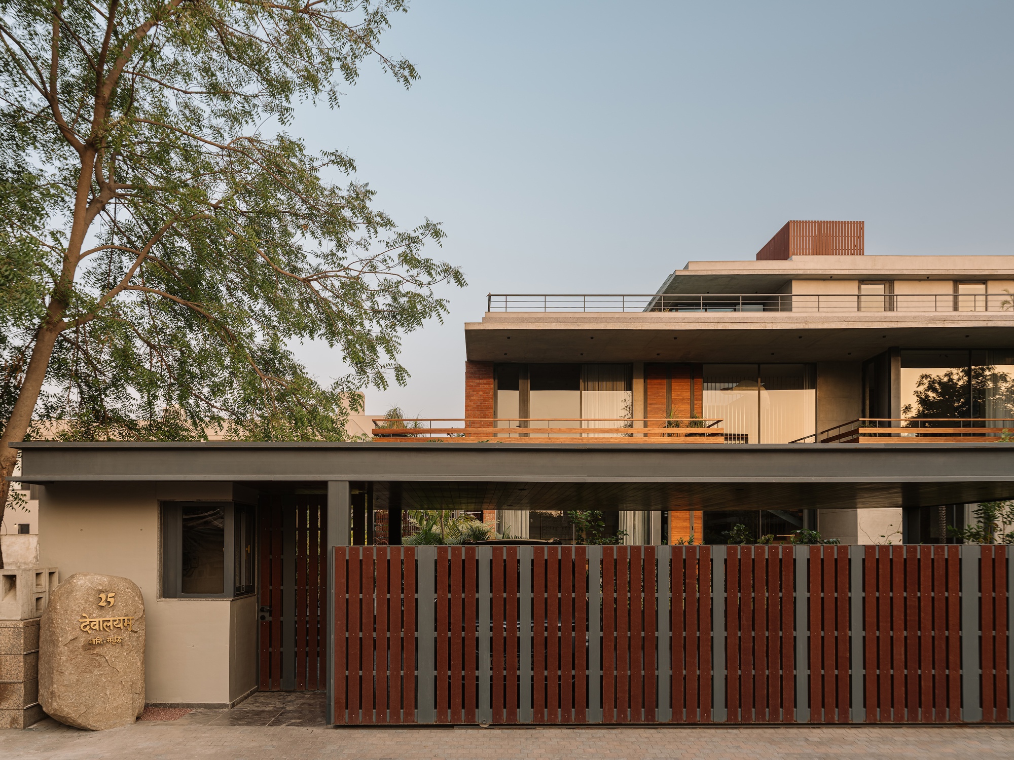 Street frontage with vertical timber slat gate, boulder nameplate and a leafy tree in front of the stacked concrete and brick house