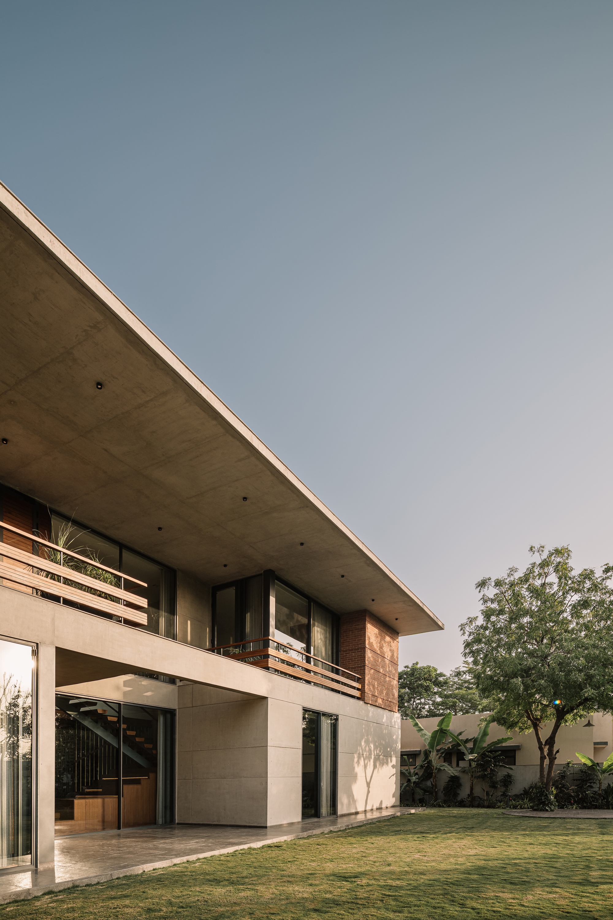 Corner of the cantilevered concrete roof slab above glazed walls and a brick panel, seen across the lawn at dusk