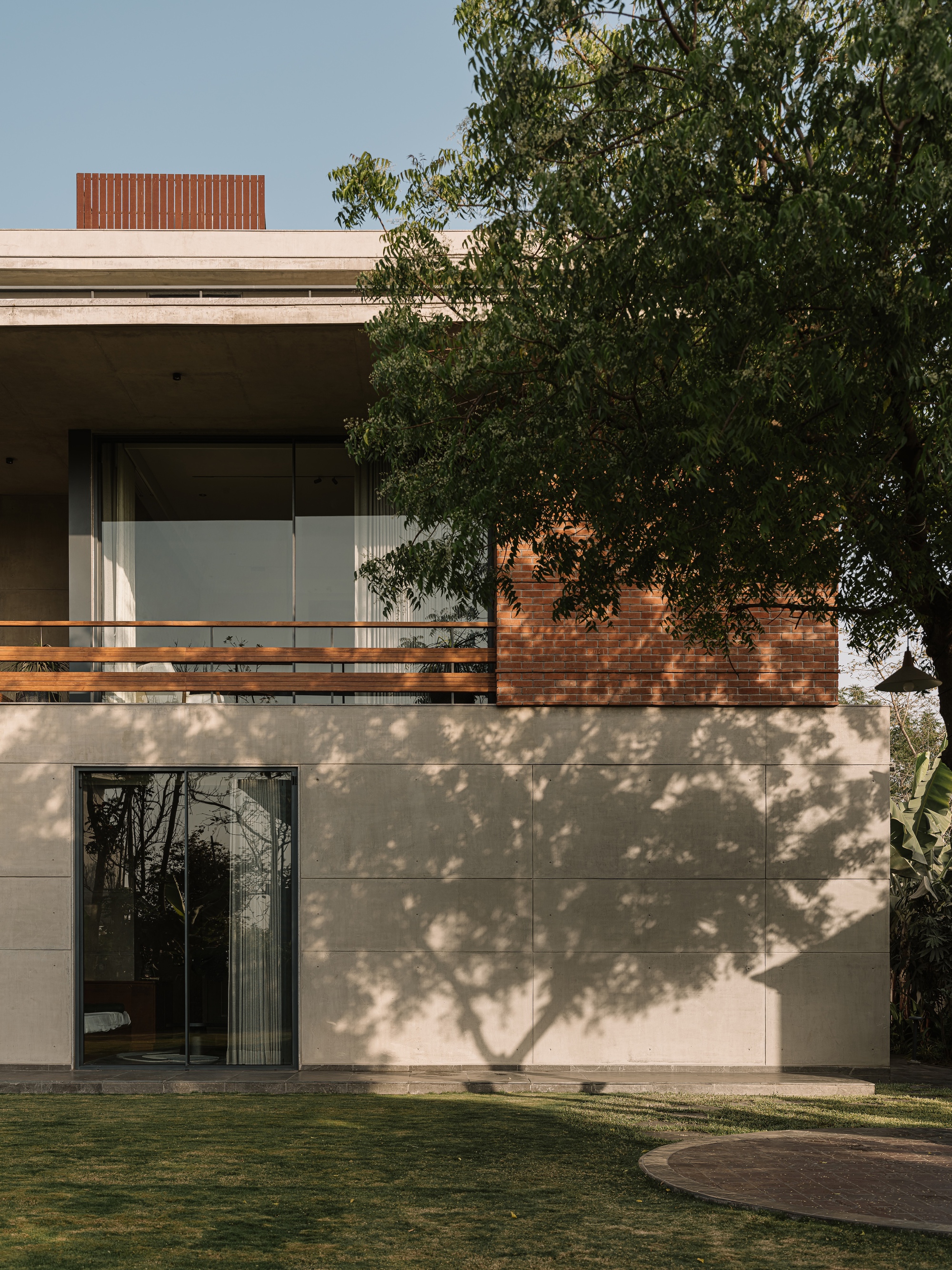 Tree shadows falling across a concrete panel wall below a brick-clad upper volume with glazing and a timber balcony rail