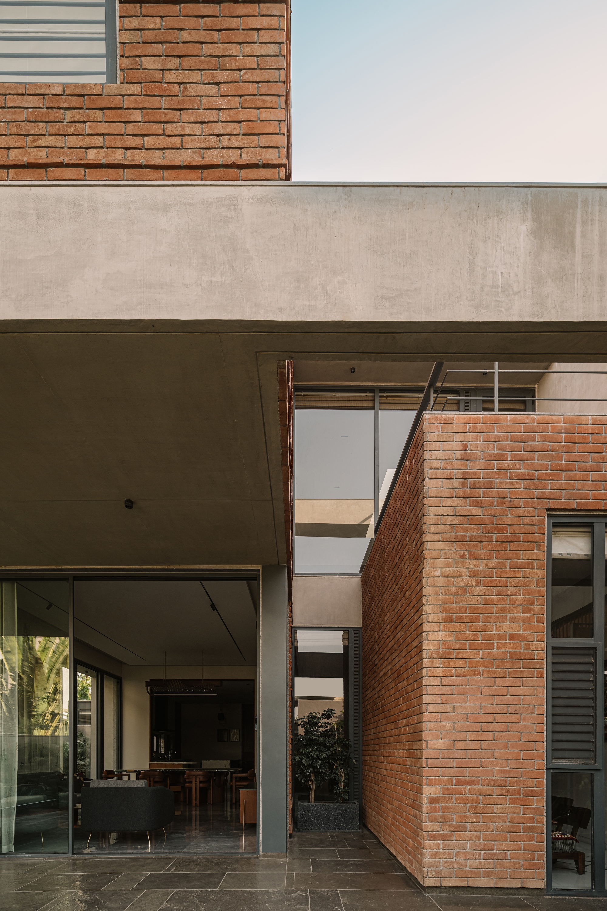 Exposed brick wall beside a concrete slab overhang and glazed living room, with slate paving in soft evening light