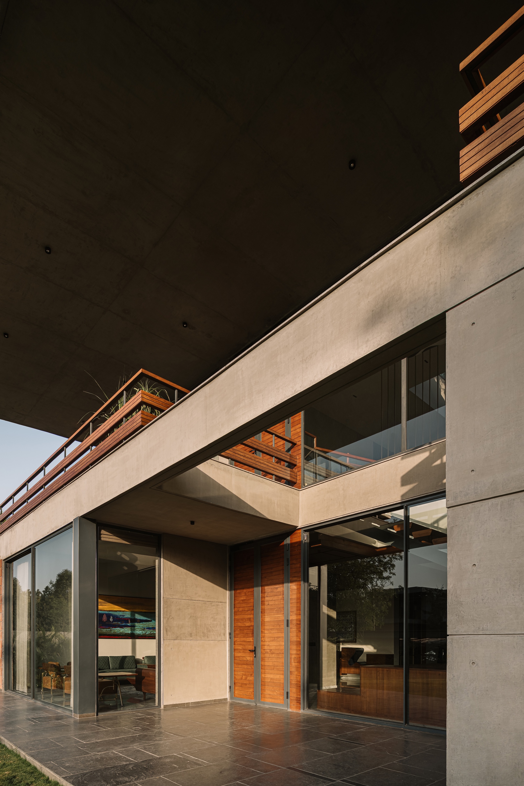Concrete-framed ground floor with timber doors and sliding glass, under a dark soffit and a planted timber balcony in late sun