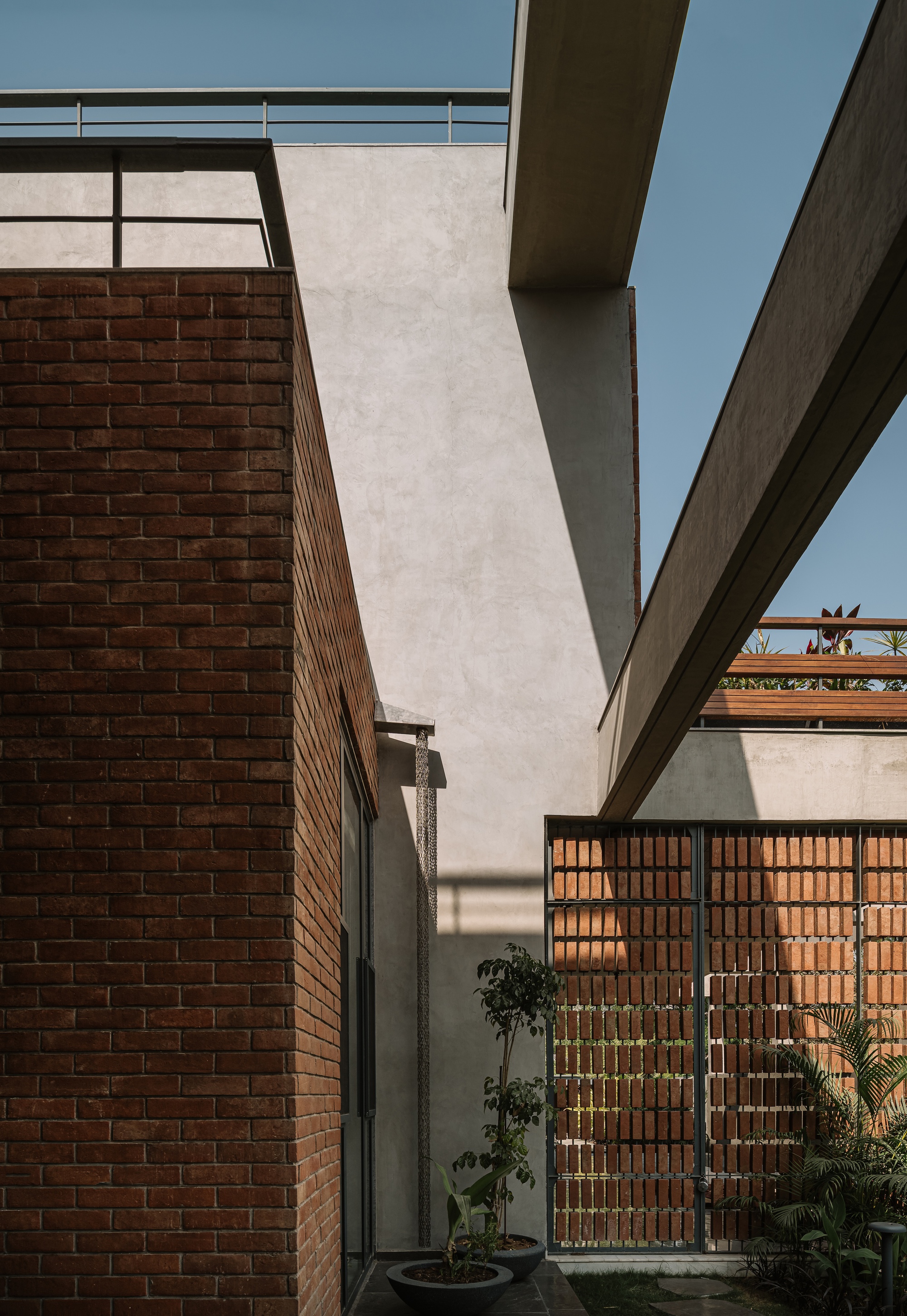 Brick corner and angled concrete beams above a perforated brick screen, with potted plants in strong midday sun under blue sky