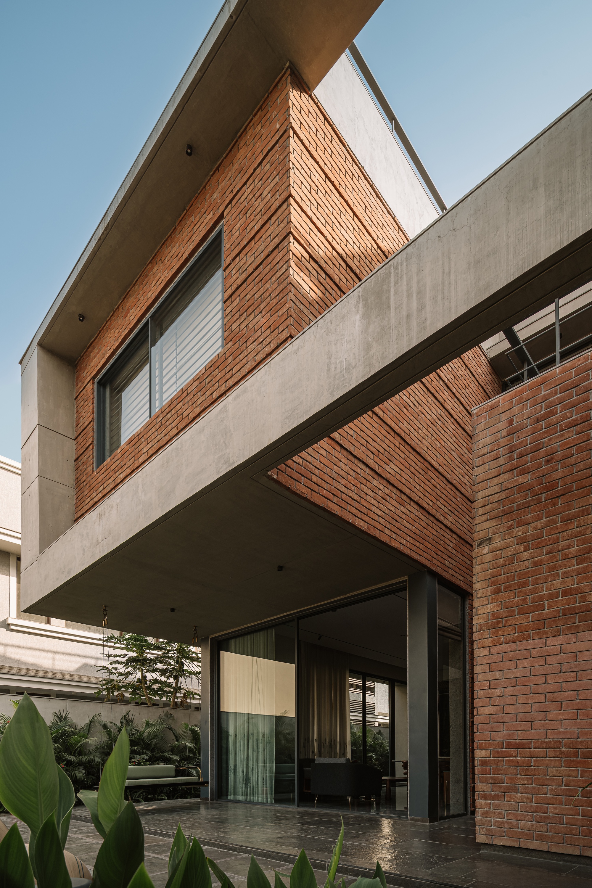 Cantilevered concrete slab over a glazed lounge, with a brick-clad upper volume and broad green leaves in the foreground