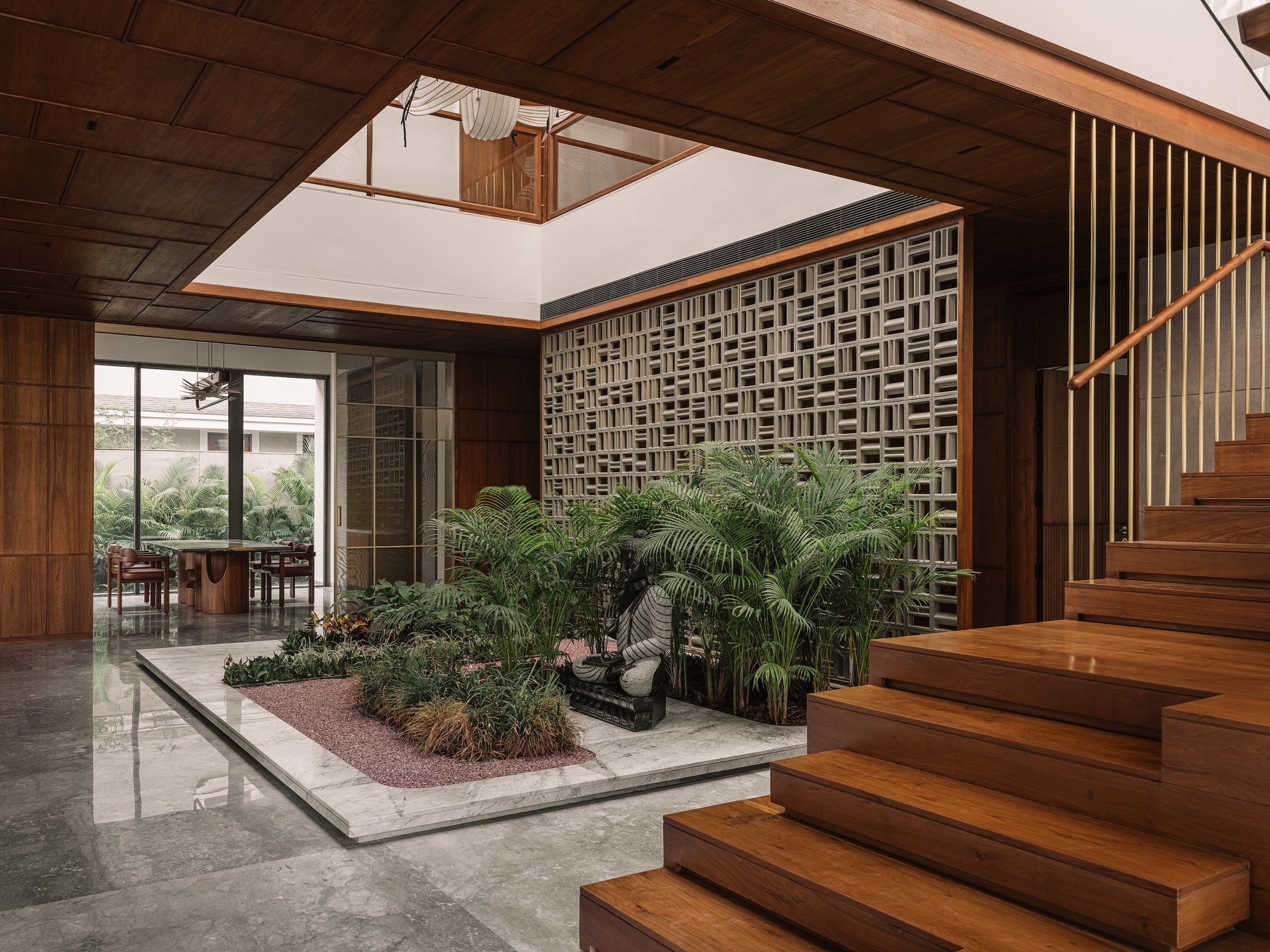 Timber stair rising beside an interior planted bed with a seated stone figure and a perforated block wall, dining area beyond