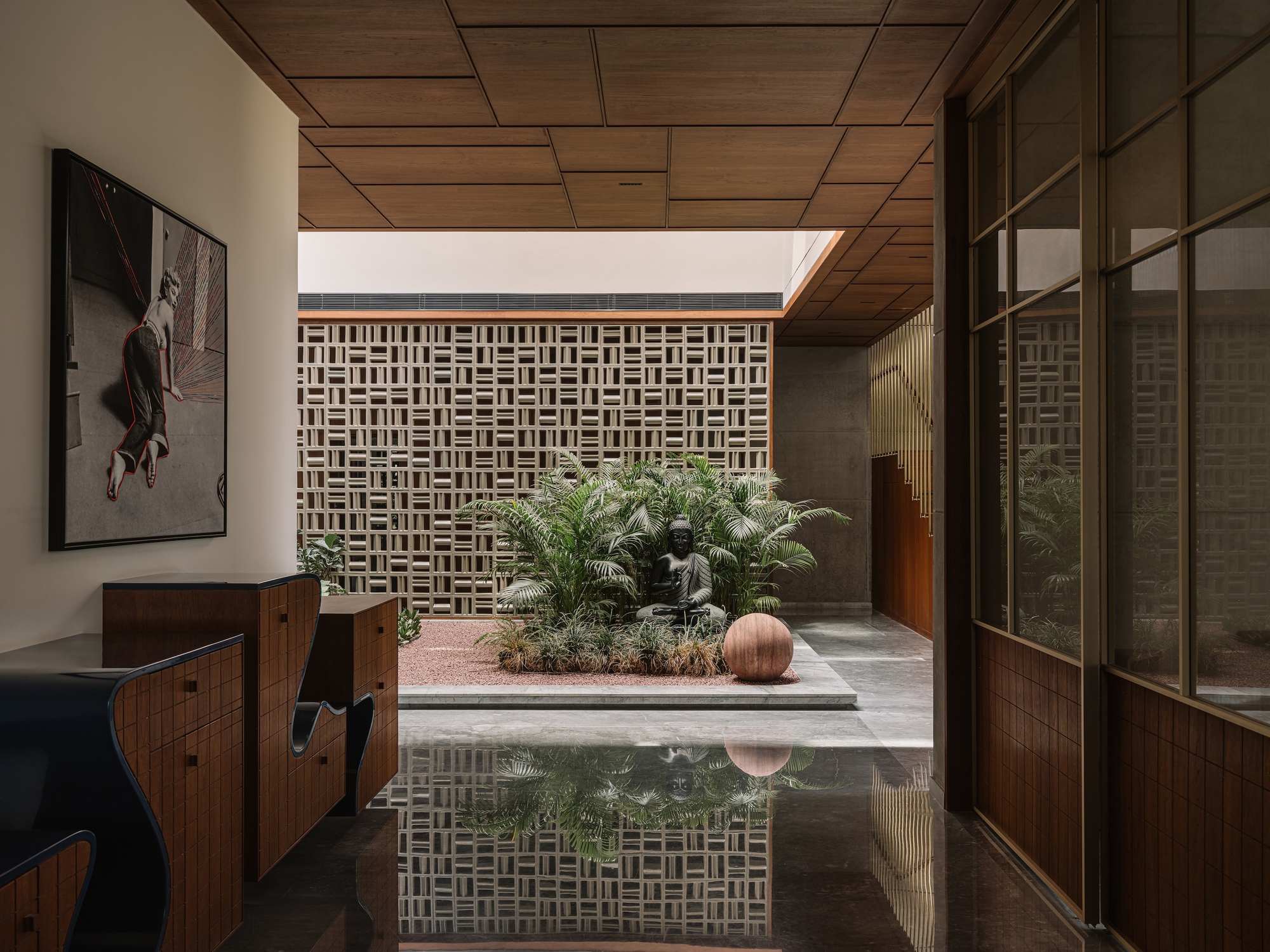 Courtyard with a seated Buddha statue, ferns and a round terracotta sphere behind a perforated brick screen, reflected in polished floor