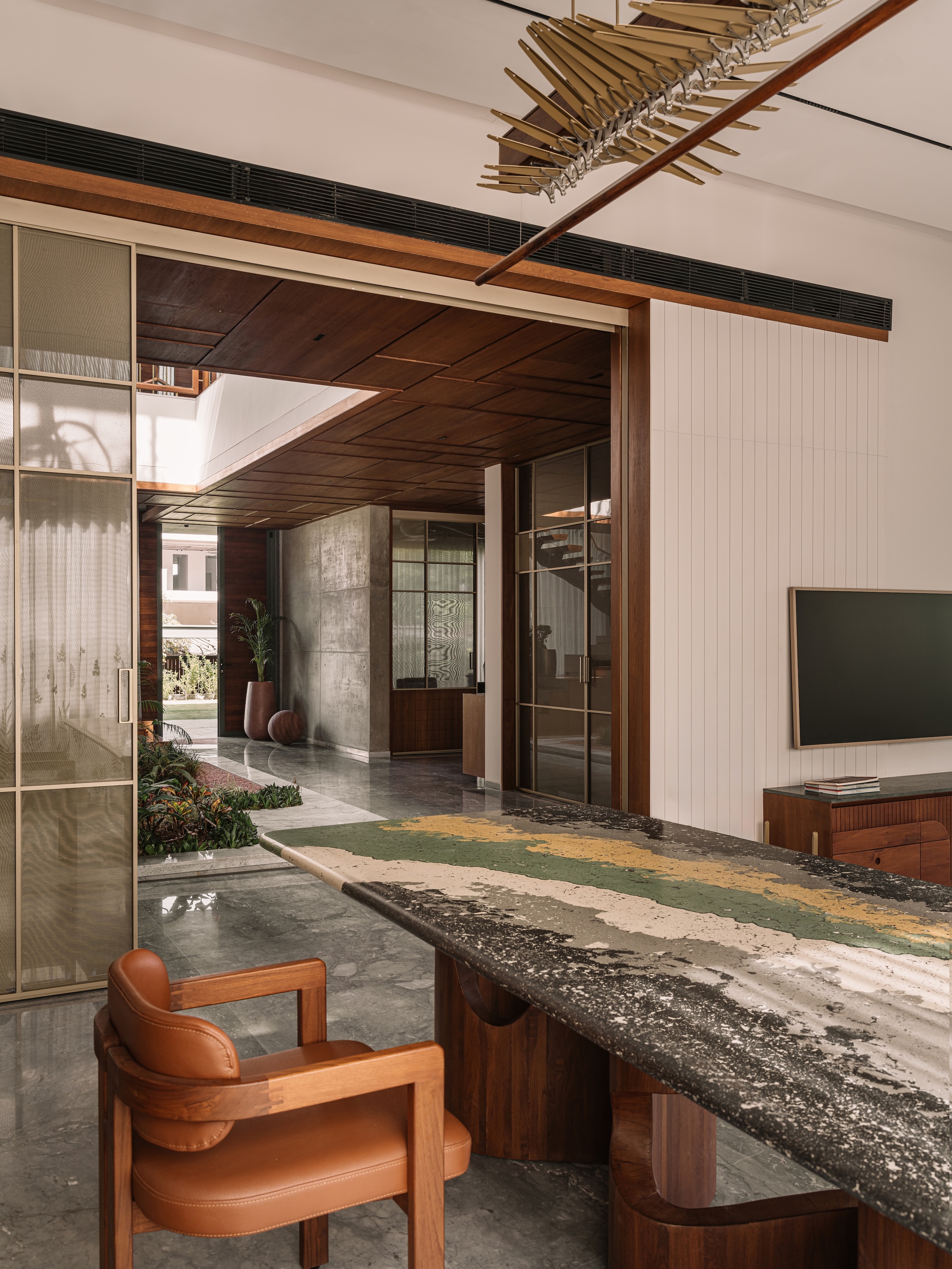 Stone-topped dining table with a leather armchair, looking through sliding glass doors toward a timber-ceilinged passage and planted courtyard