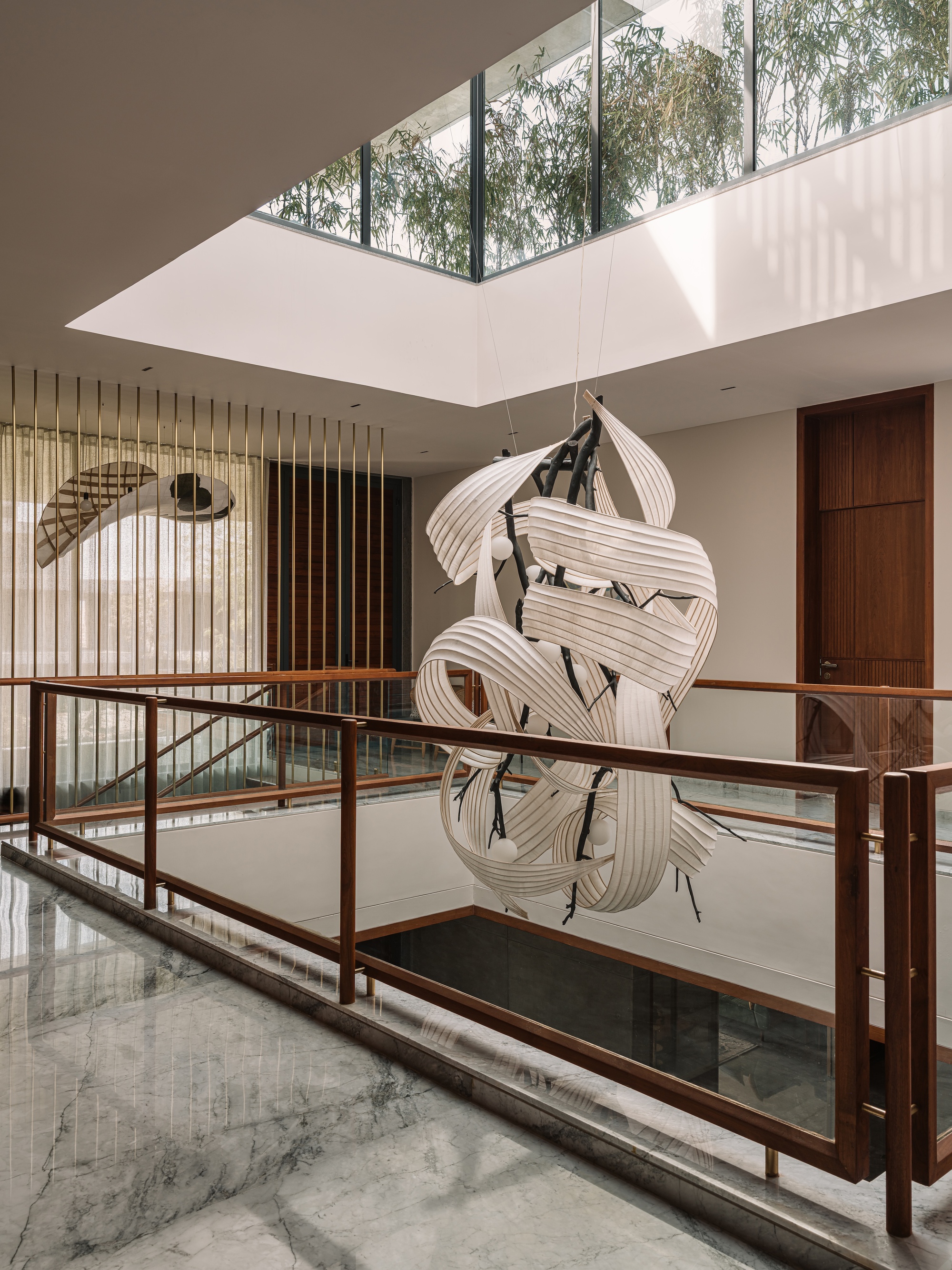 Sculptural white ribbon light fixture hanging above a glass-and-timber balustrade beneath a skylight filled with bamboo foliage