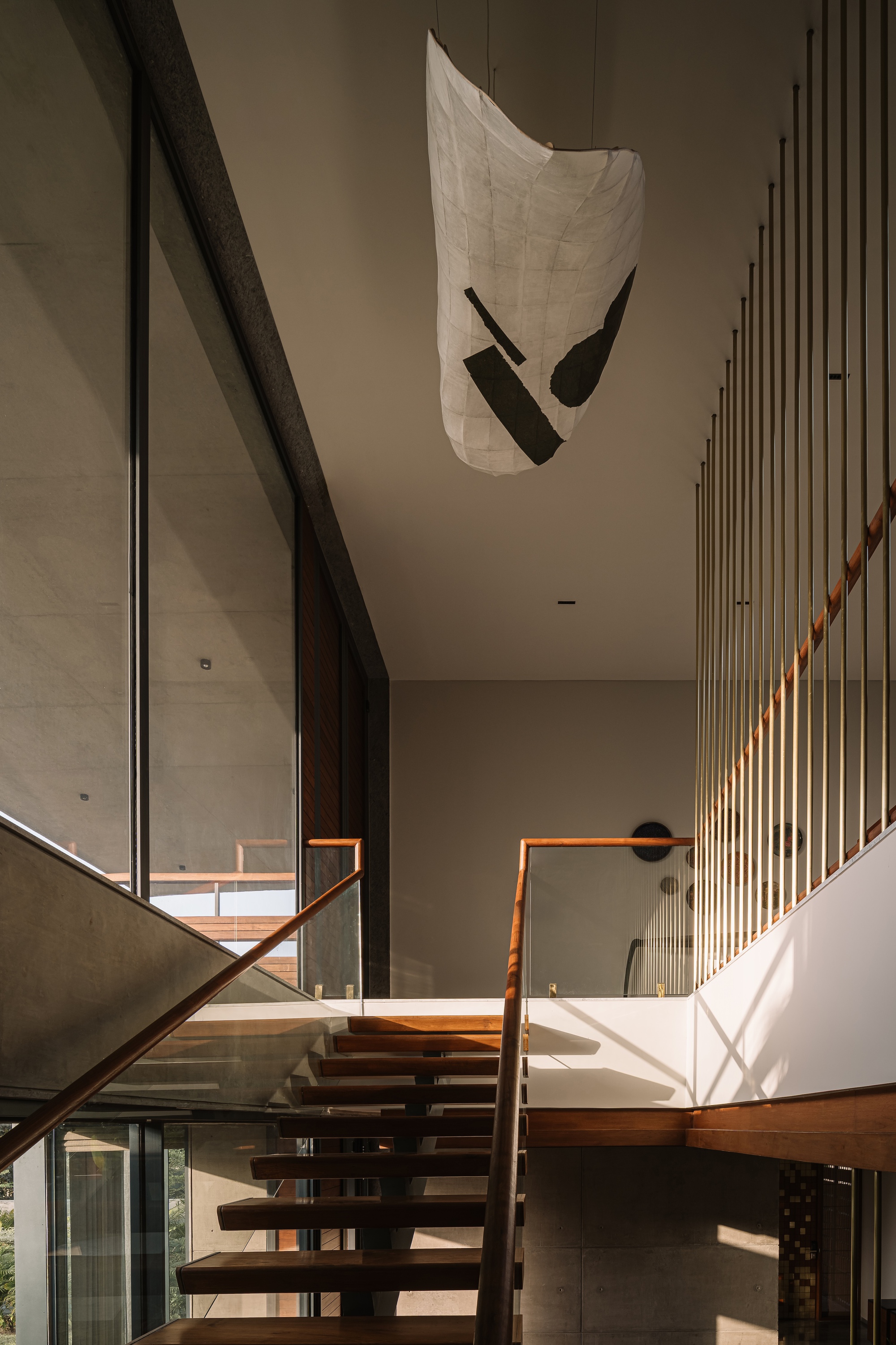 Open-tread timber staircase with a glass balustrade and a paper lantern hanging beside vertical brass rods in raking sunlight