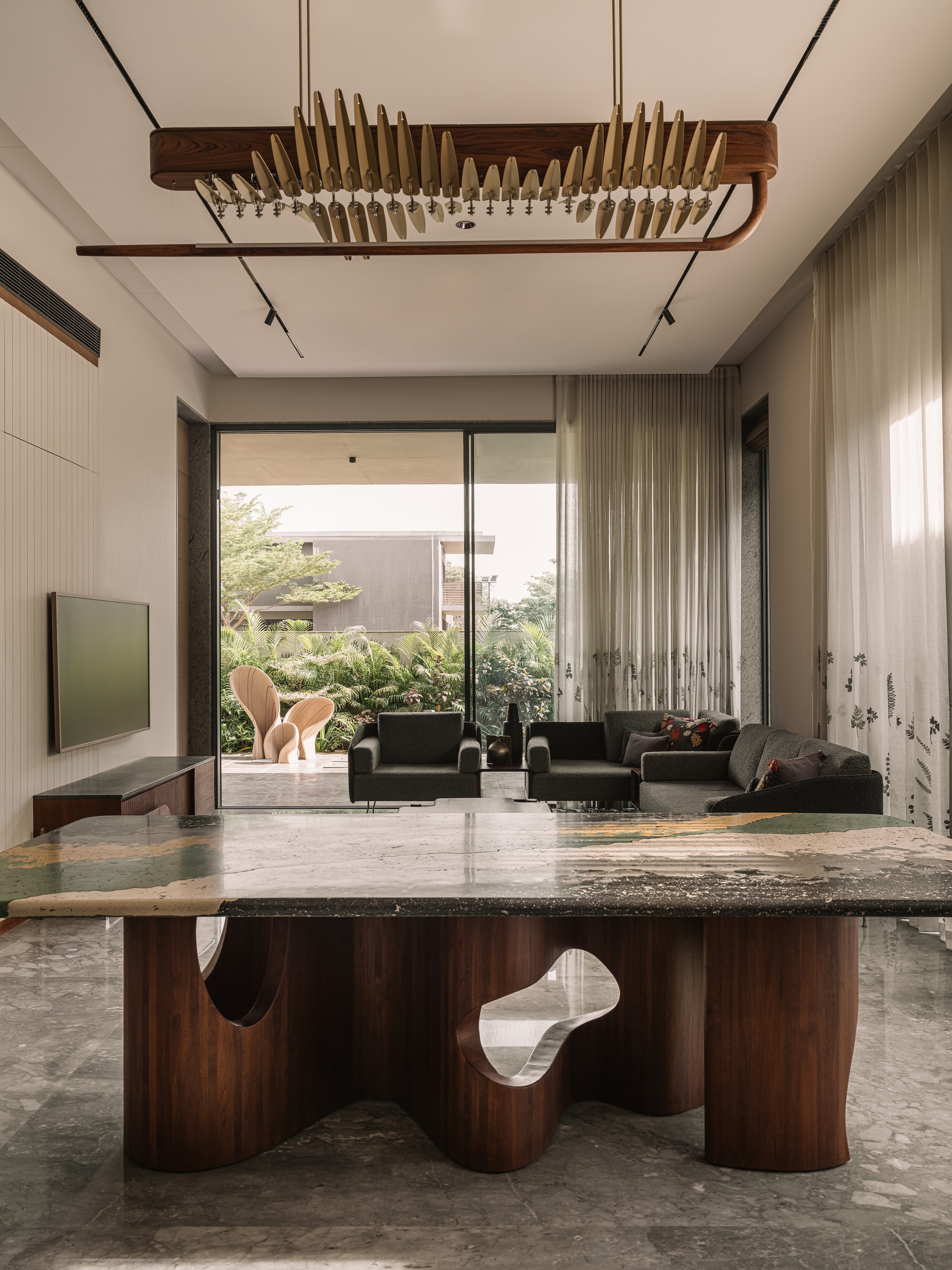 Dining table with a sculpted walnut base and stone top facing a lounge, sheer curtains and a garden through sliding glass