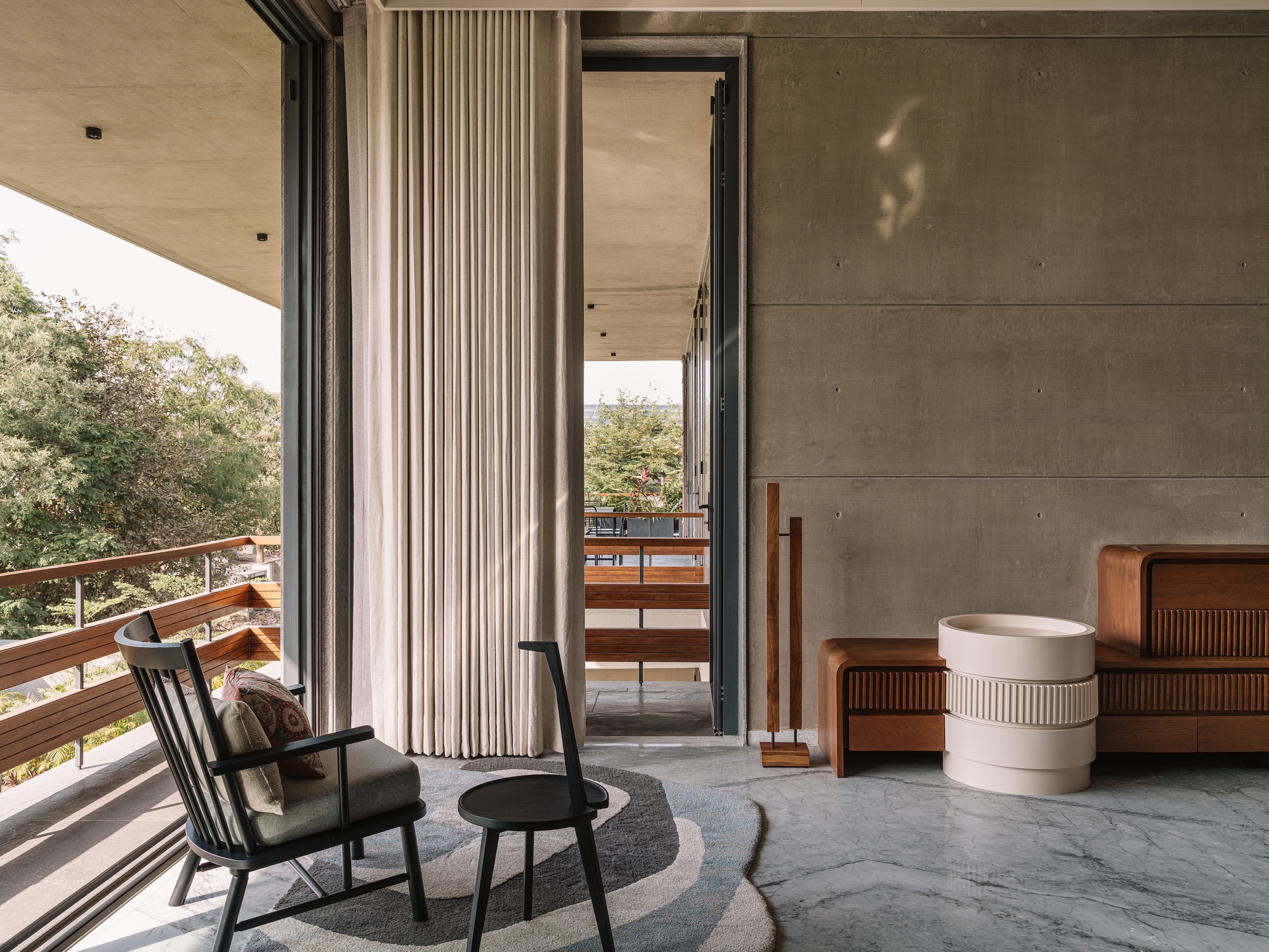 Balcony lounge with a black rocking chair, pleated linen curtain and board-marked concrete wall in warm afternoon light