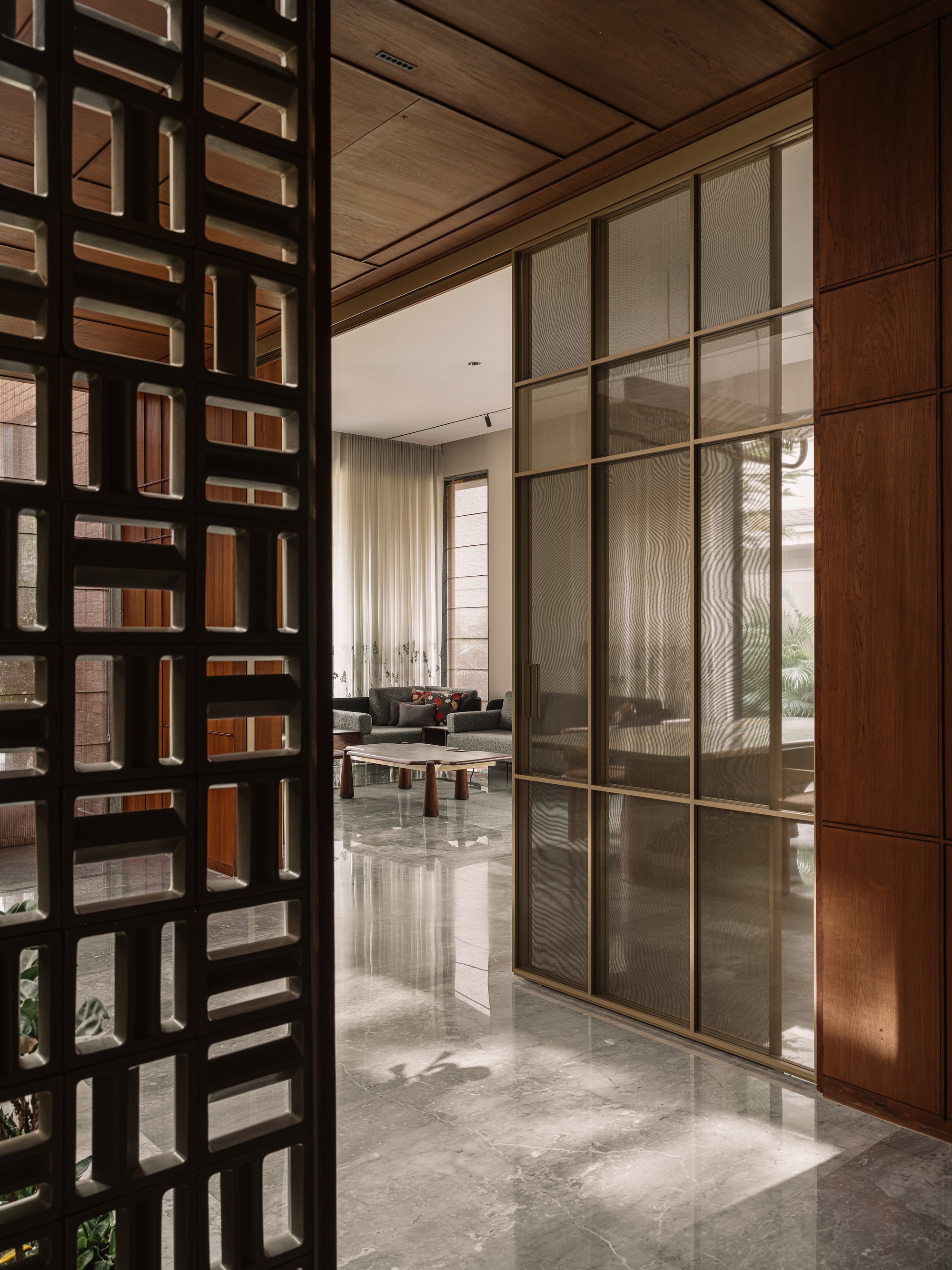 Perforated screen wall in the foreground, with a brass-framed fluted glass partition leading to a sitting area, sunlight on polished stone floor