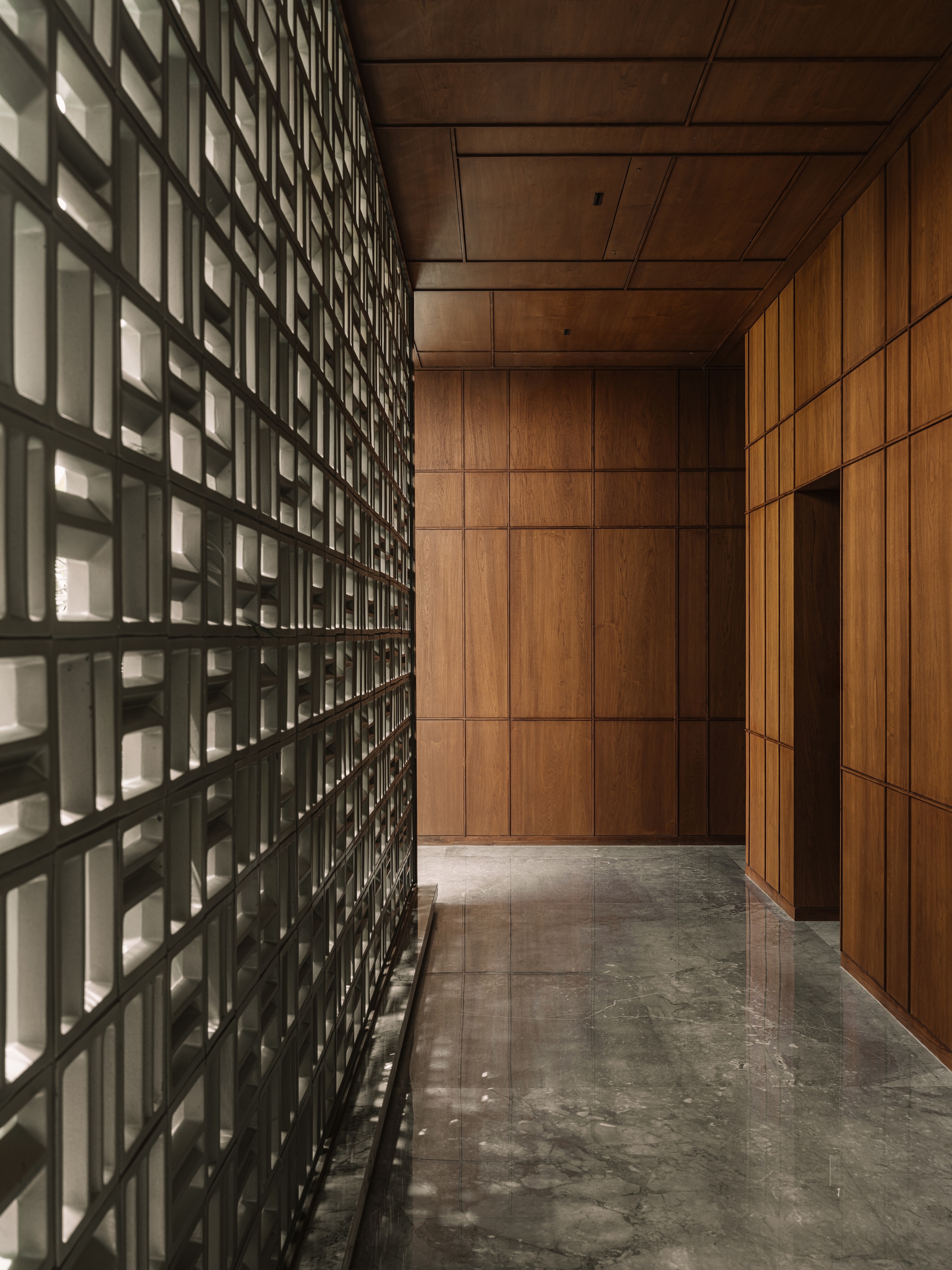 Corridor lined with a white ceramic block lattice wall opposite walnut paneling, with a coffered timber ceiling above grey marble