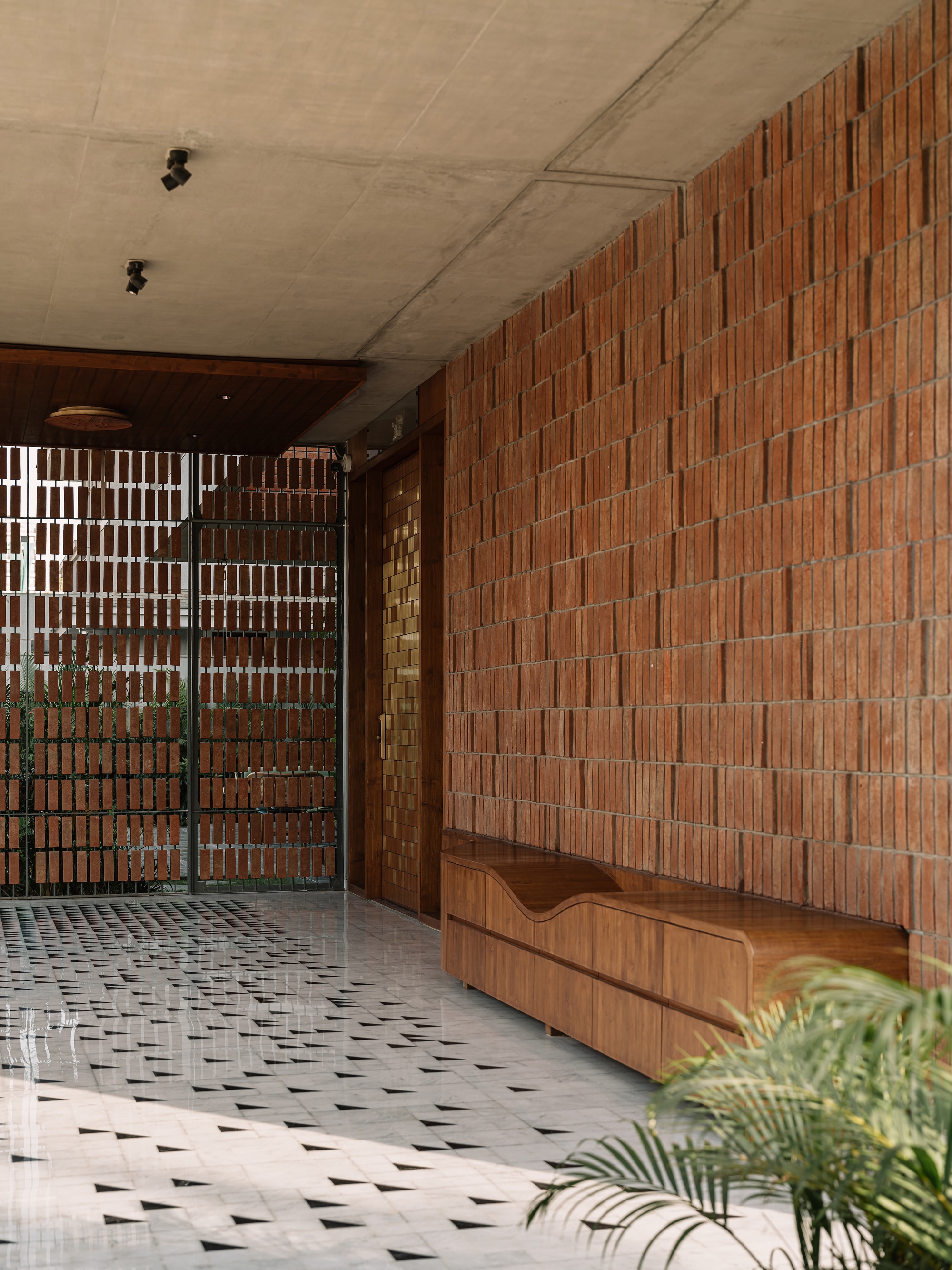 Covered entry with a vertical brick wall, built-in timber bench and triangle-patterned tile floor, a palm frond in the corner