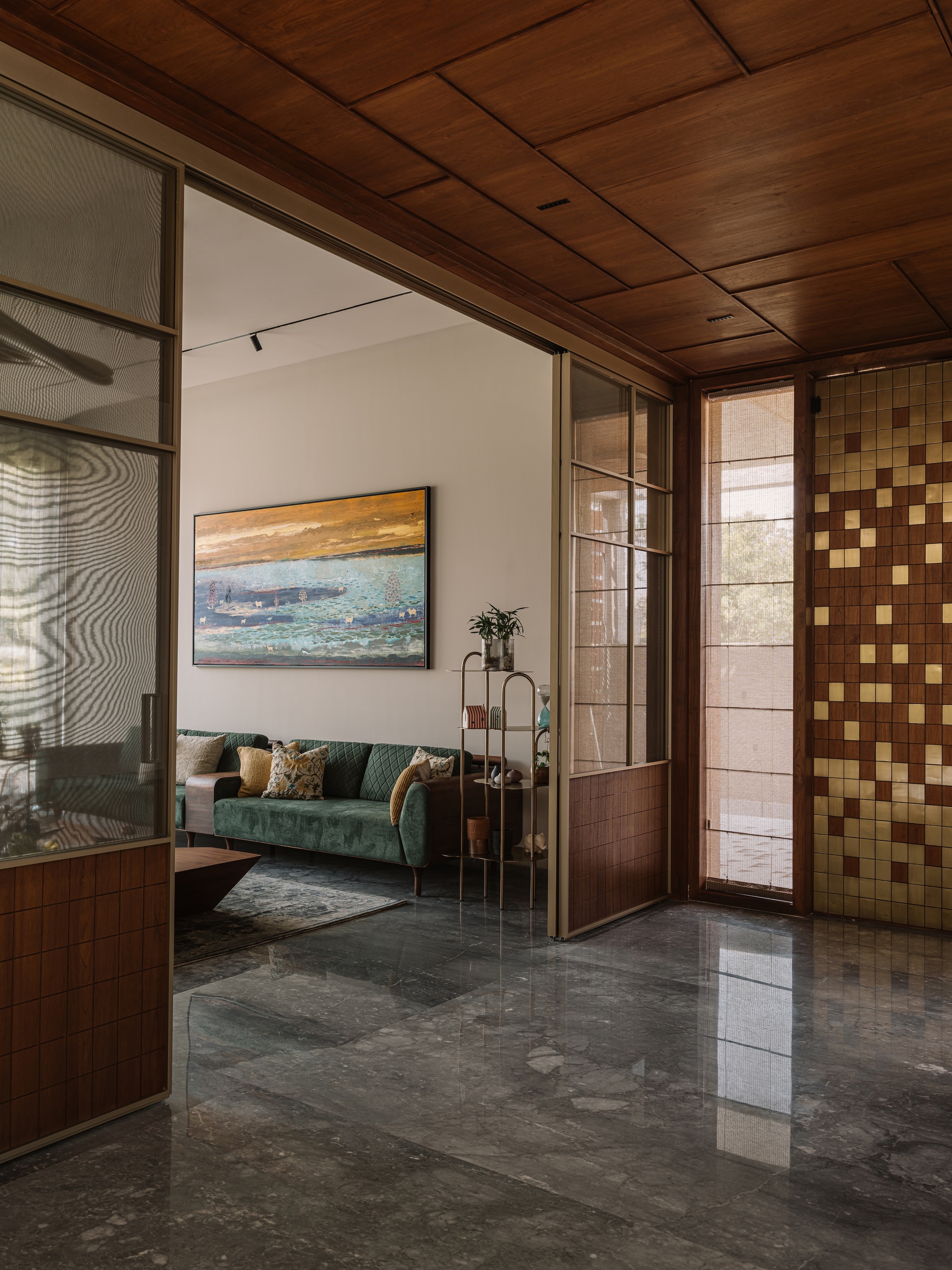 Entry area with a timber ceiling, glazed sliding partitions and gold mosaic tile wall opening onto a green sofa and painting