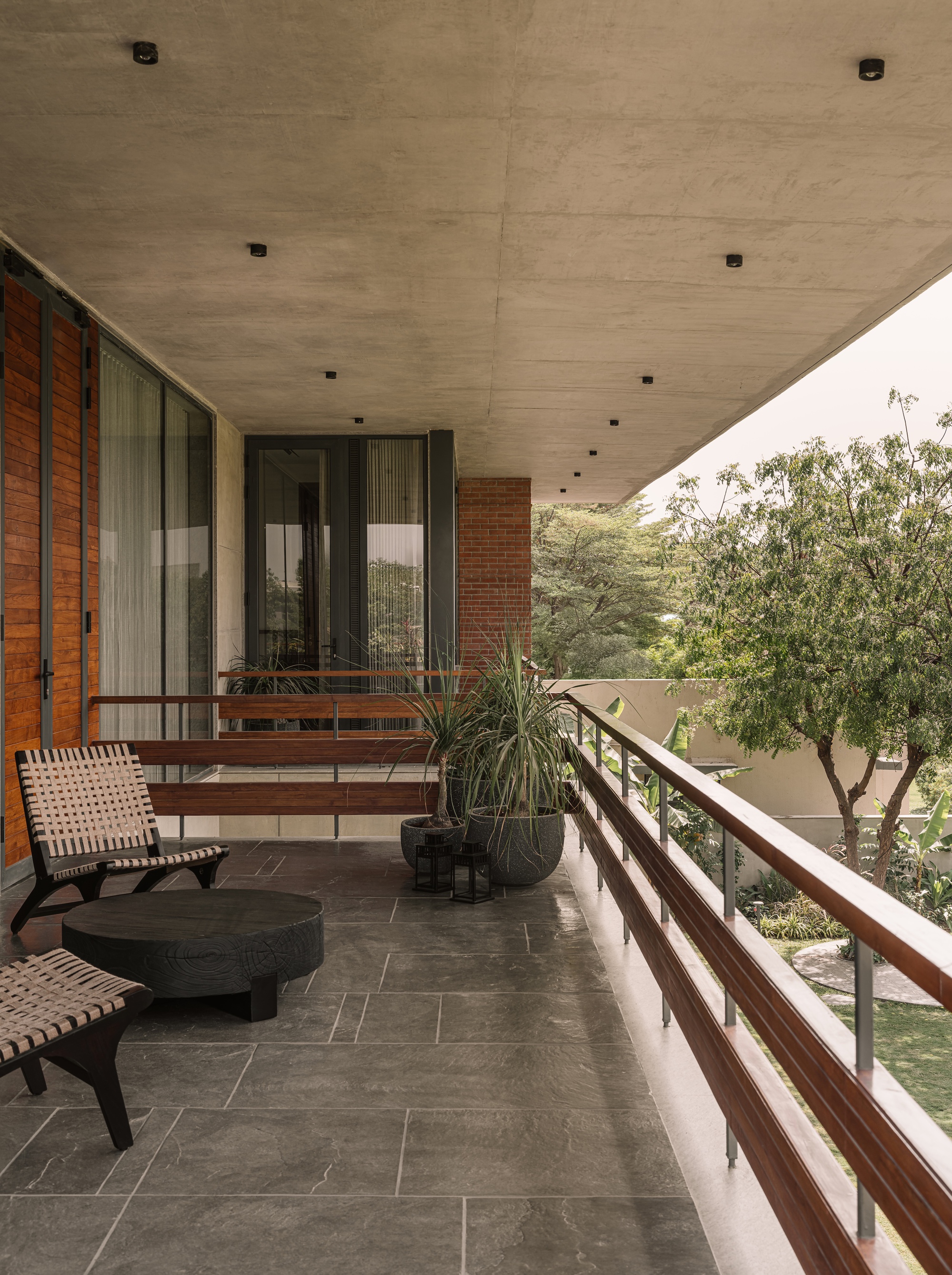Covered balcony with a concrete soffit, timber railing, woven lounge chairs and potted plants facing a tree in daylight