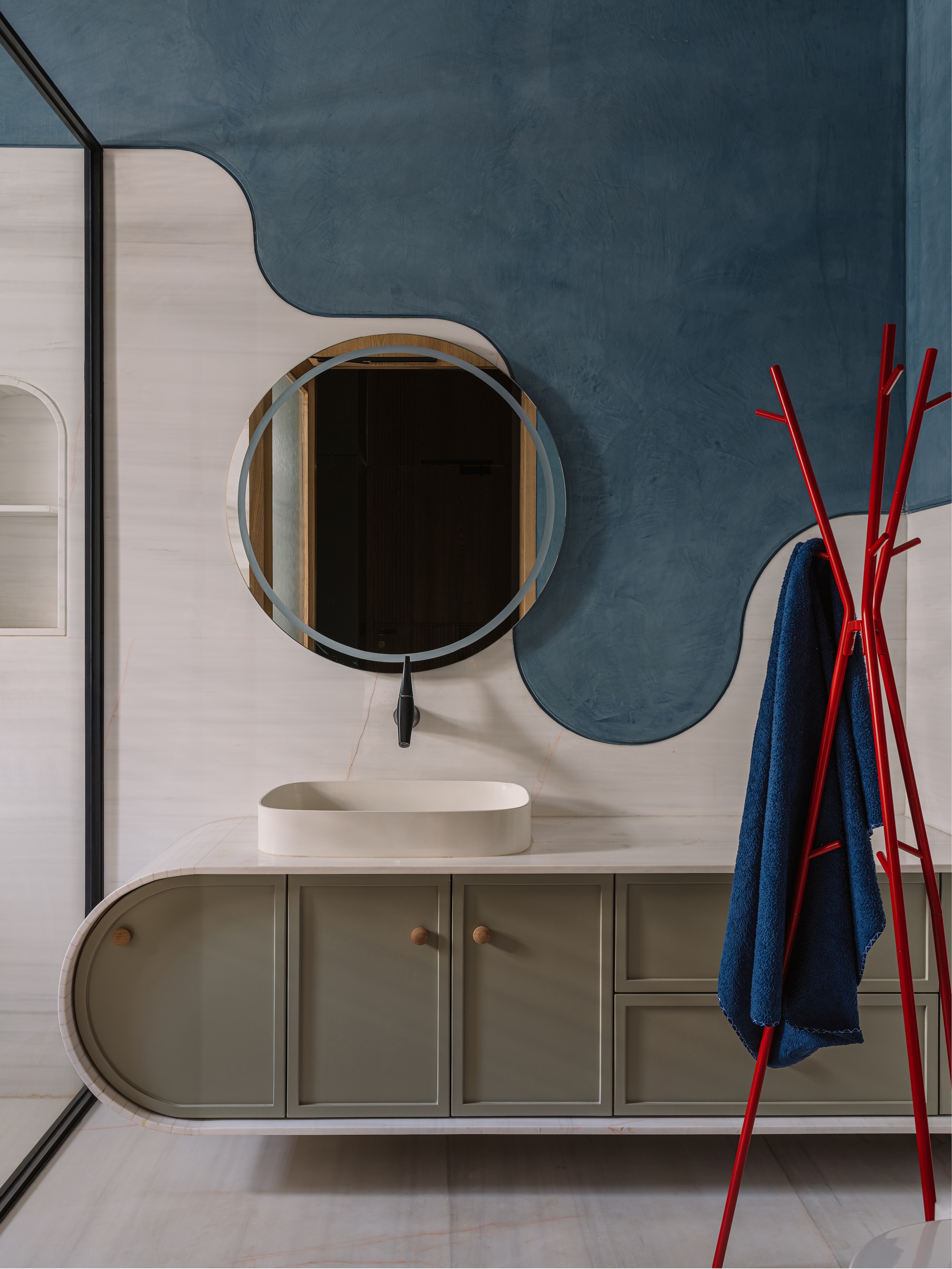 Floating curved vanity with a round mirror against a wavy blue plaster wall and a red coat stand holding a towel