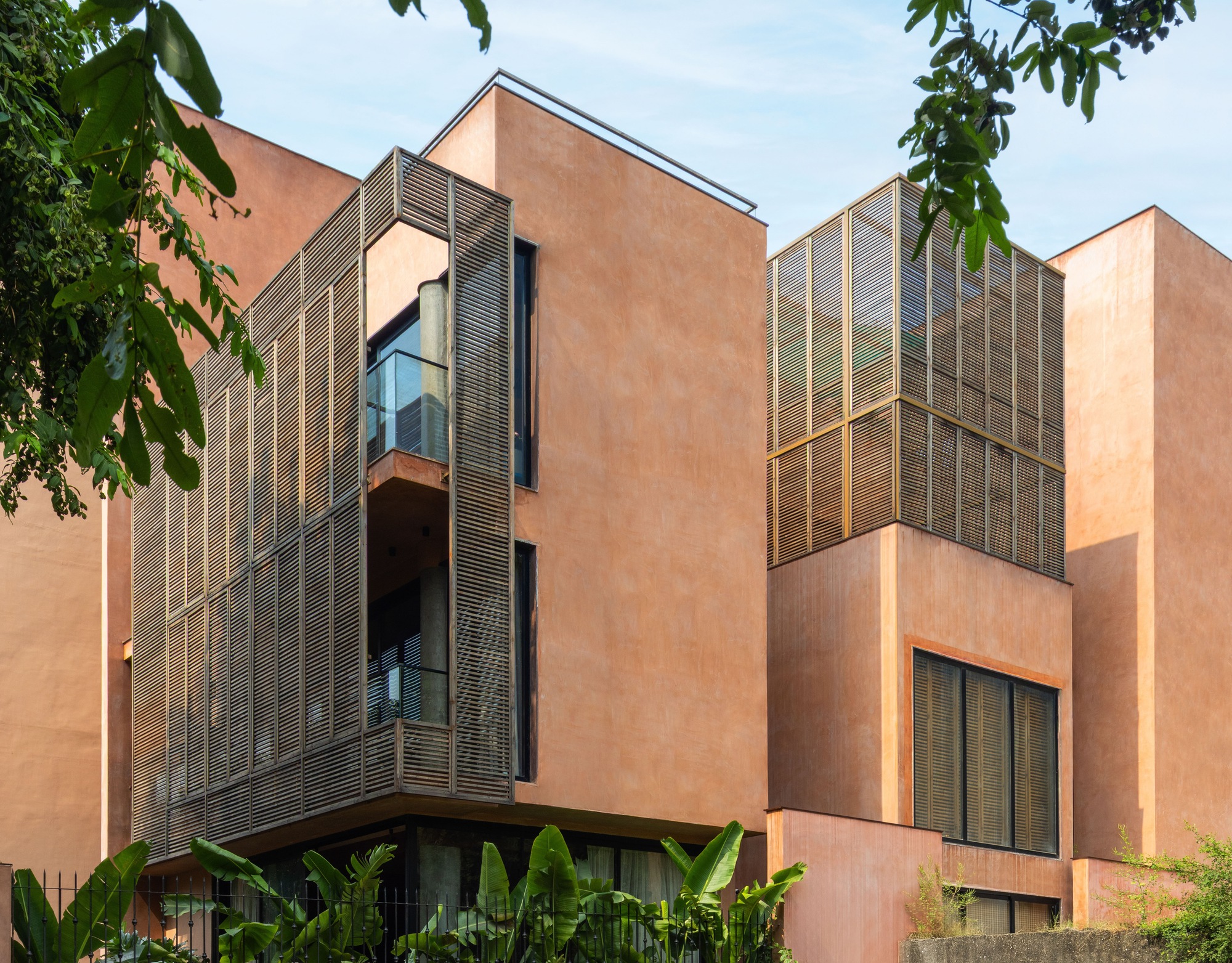 Terracotta plastered facade with timber louvre screens wrapping a corner balcony, framed by leafy branches under a pale blue sky