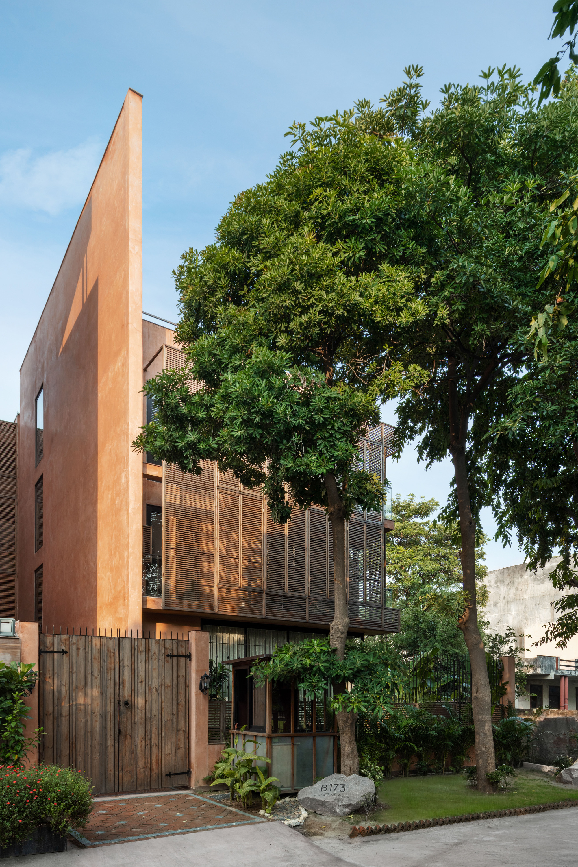 Street view of a tall blade wall and timber louvred upper floors behind mature trees and a plank gate