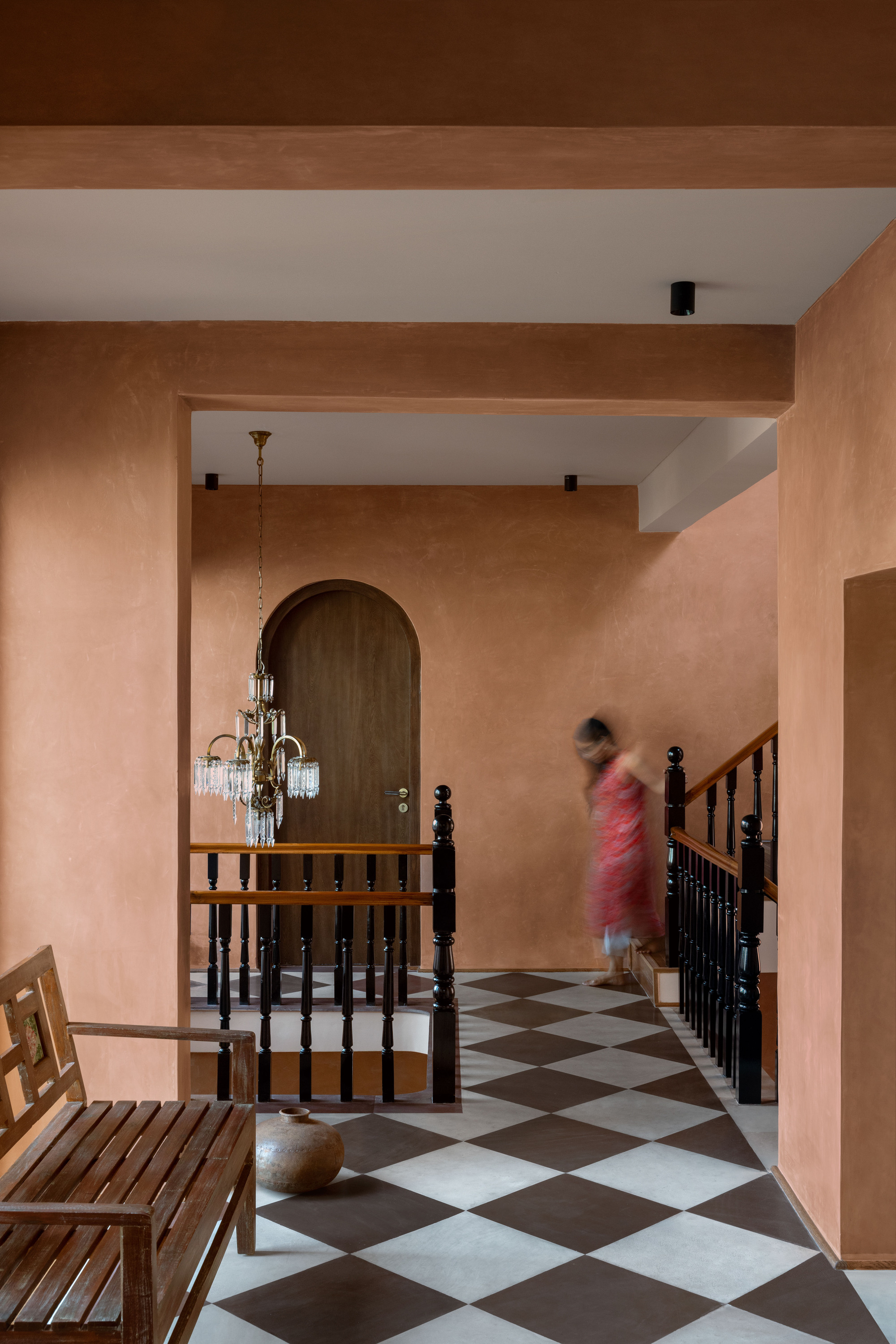 Landing with chequerboard floor, black balustrade and arched timber door, a blurred figure descending the stairs beside a crystal chandelier