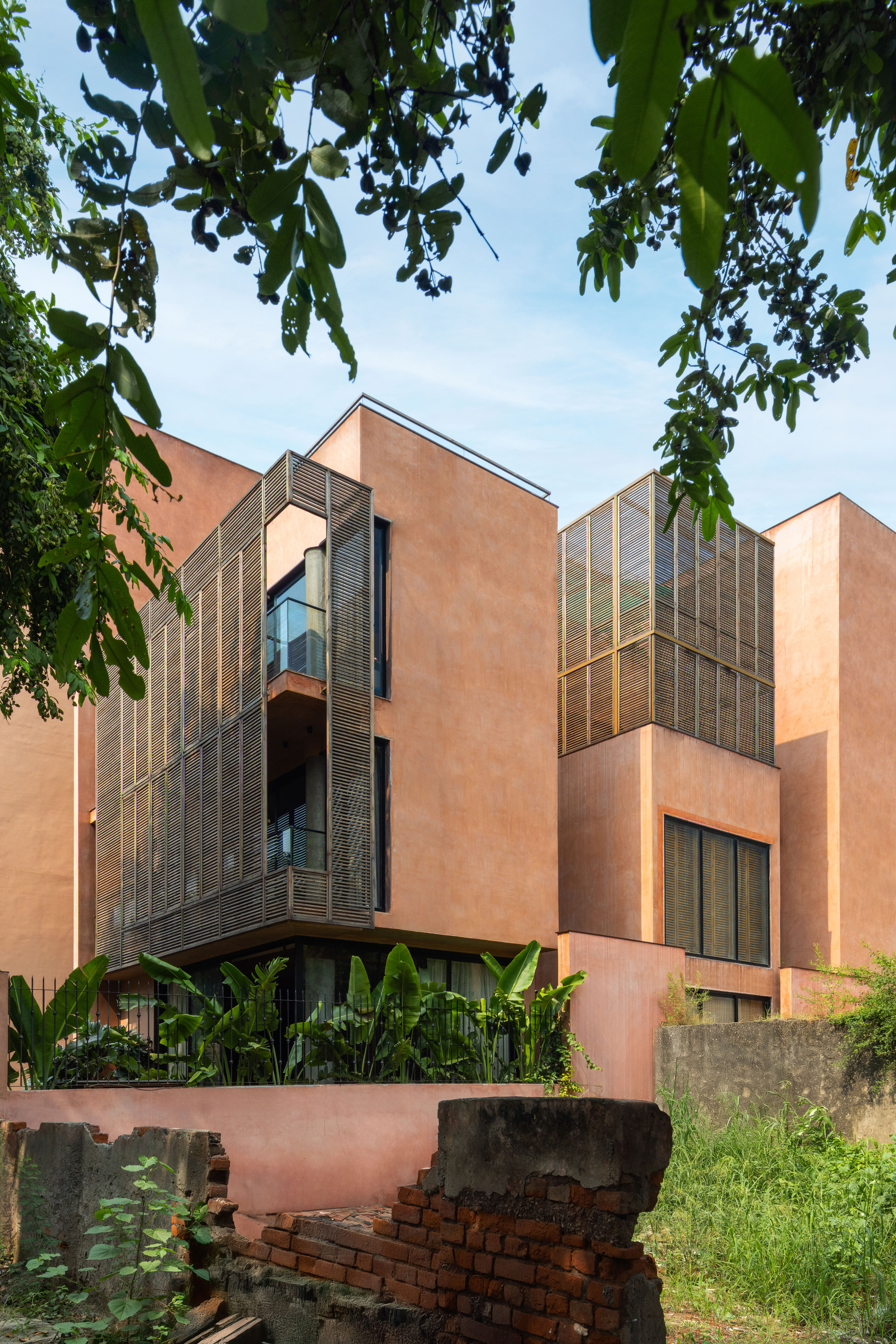 Terracotta-plastered stepped volumes with timber louvre screens rising behind a ruined brick wall and banana plants, framed by leaves against blue sky