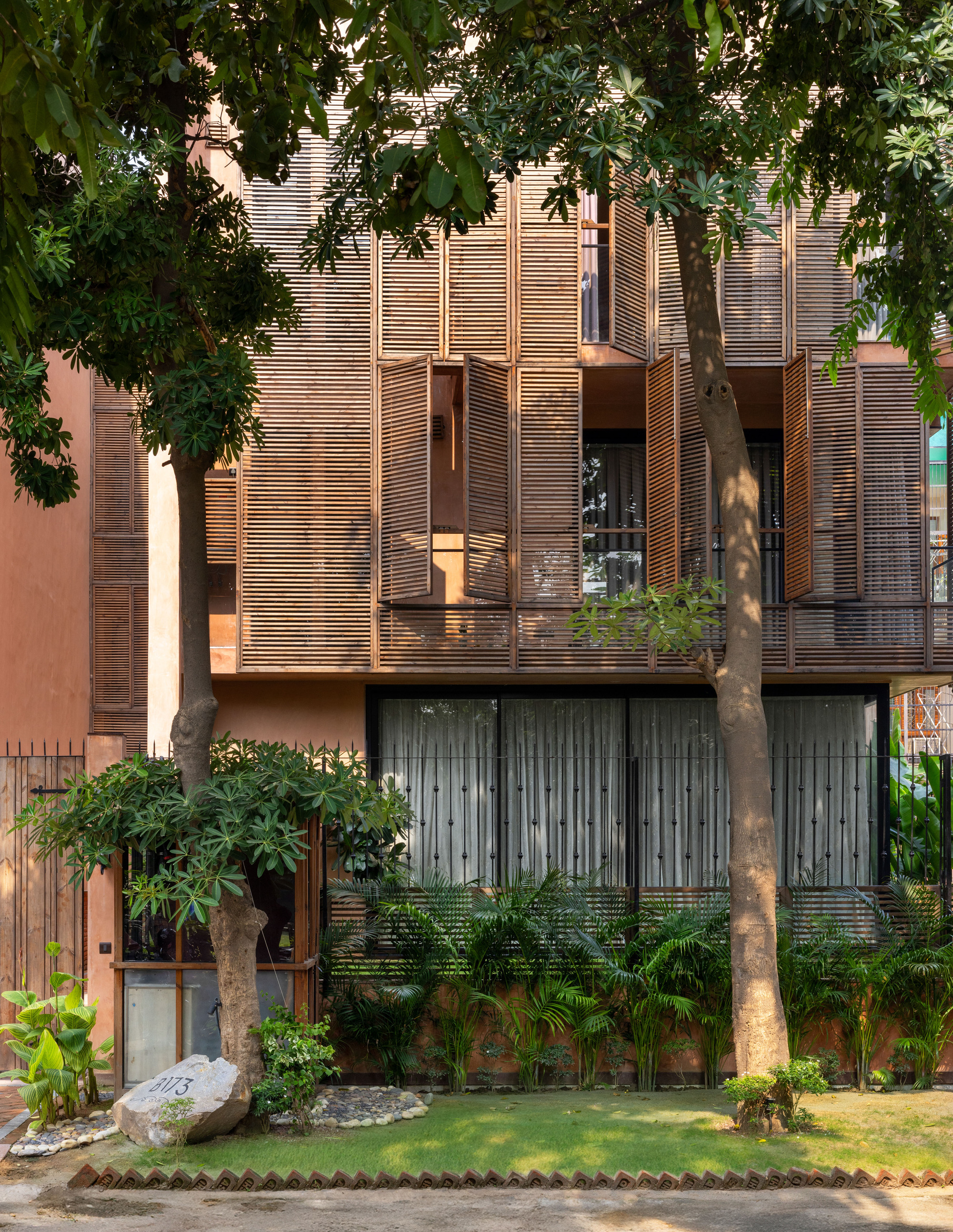 Street facade with open timber louvre shutters above sheer curtains, seen through two tree trunks and a planted garden in daylight