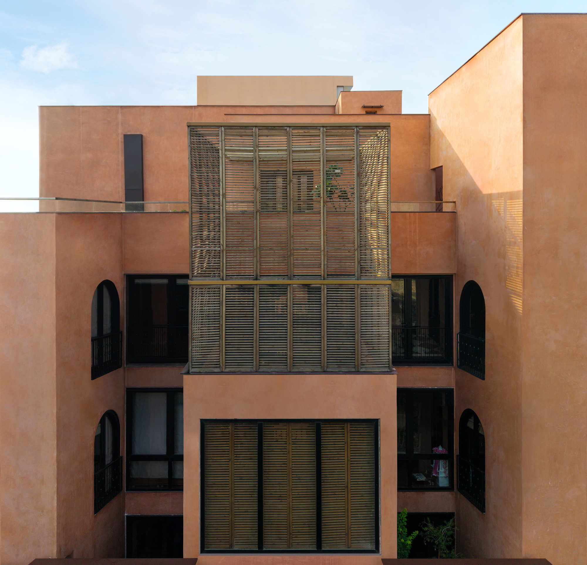 Rear elevation of ochre plastered volumes with arched windows and a central louvred timber box under a pale afternoon sky