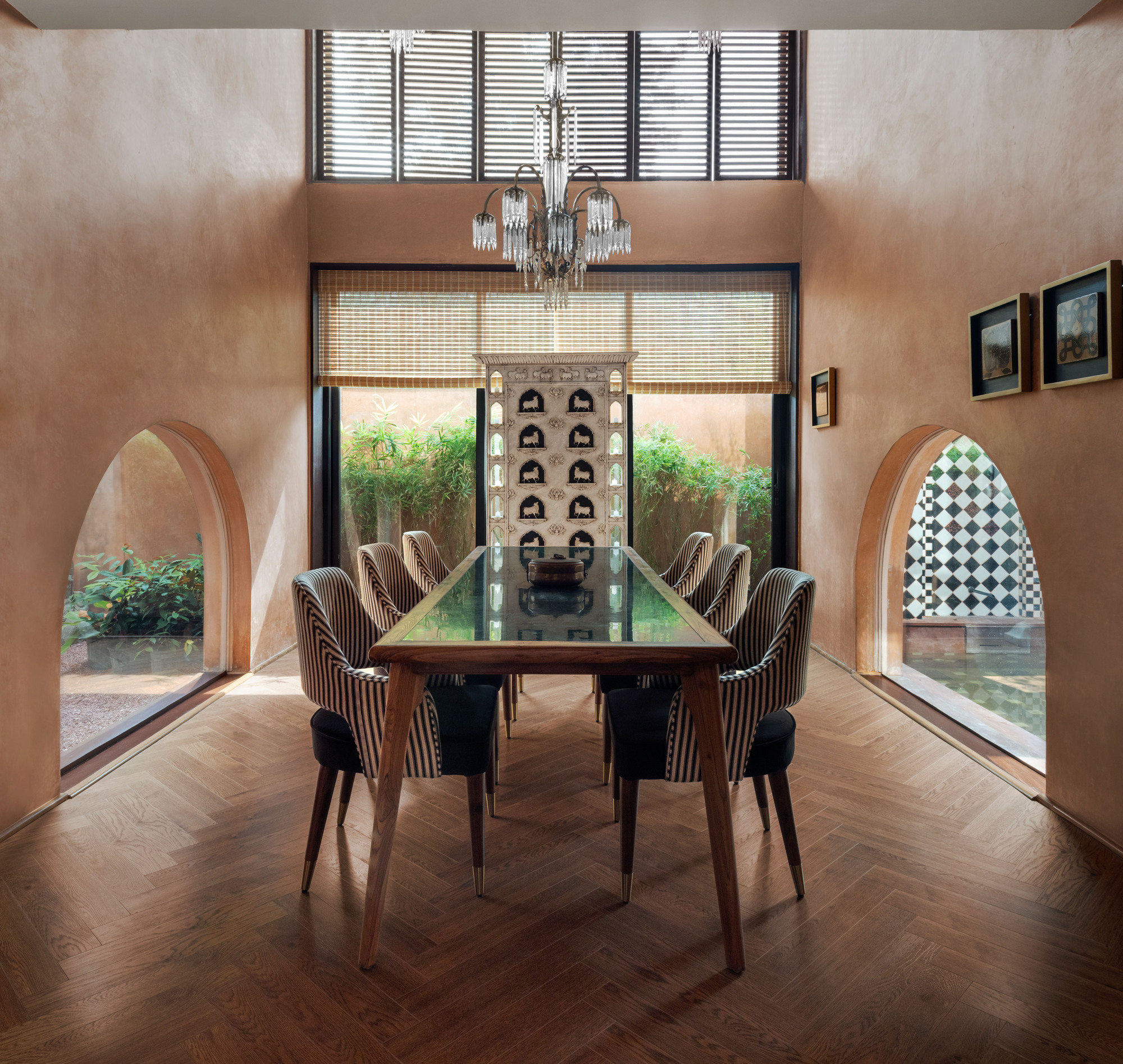 Double-height dining room with striped chairs, glass-topped table and crystal chandelier between two arched openings onto garden courtyards