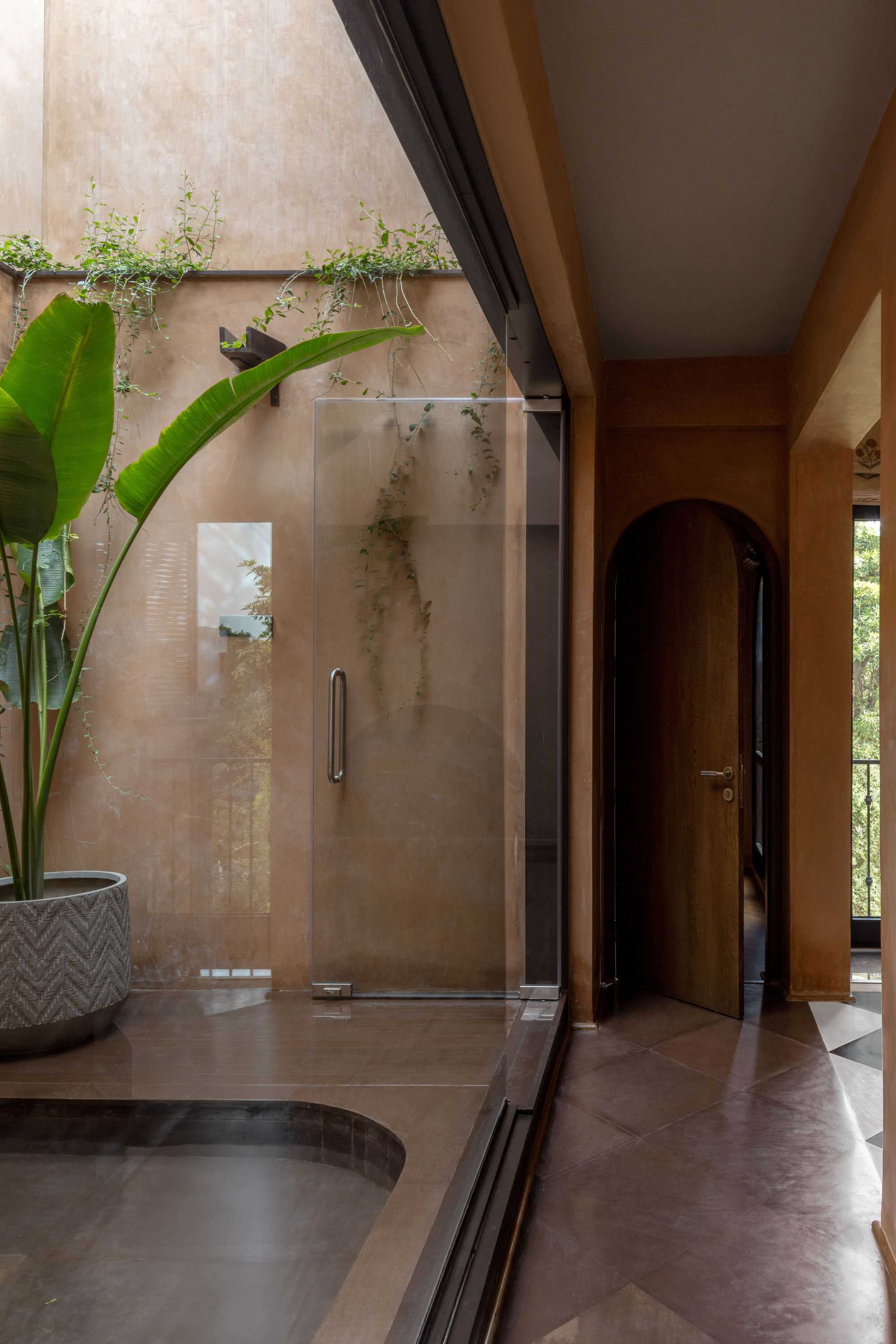 Glass door beside a sunken bath, with a potted strelitzia, trailing vines and an arched timber door beyond in soft daylight