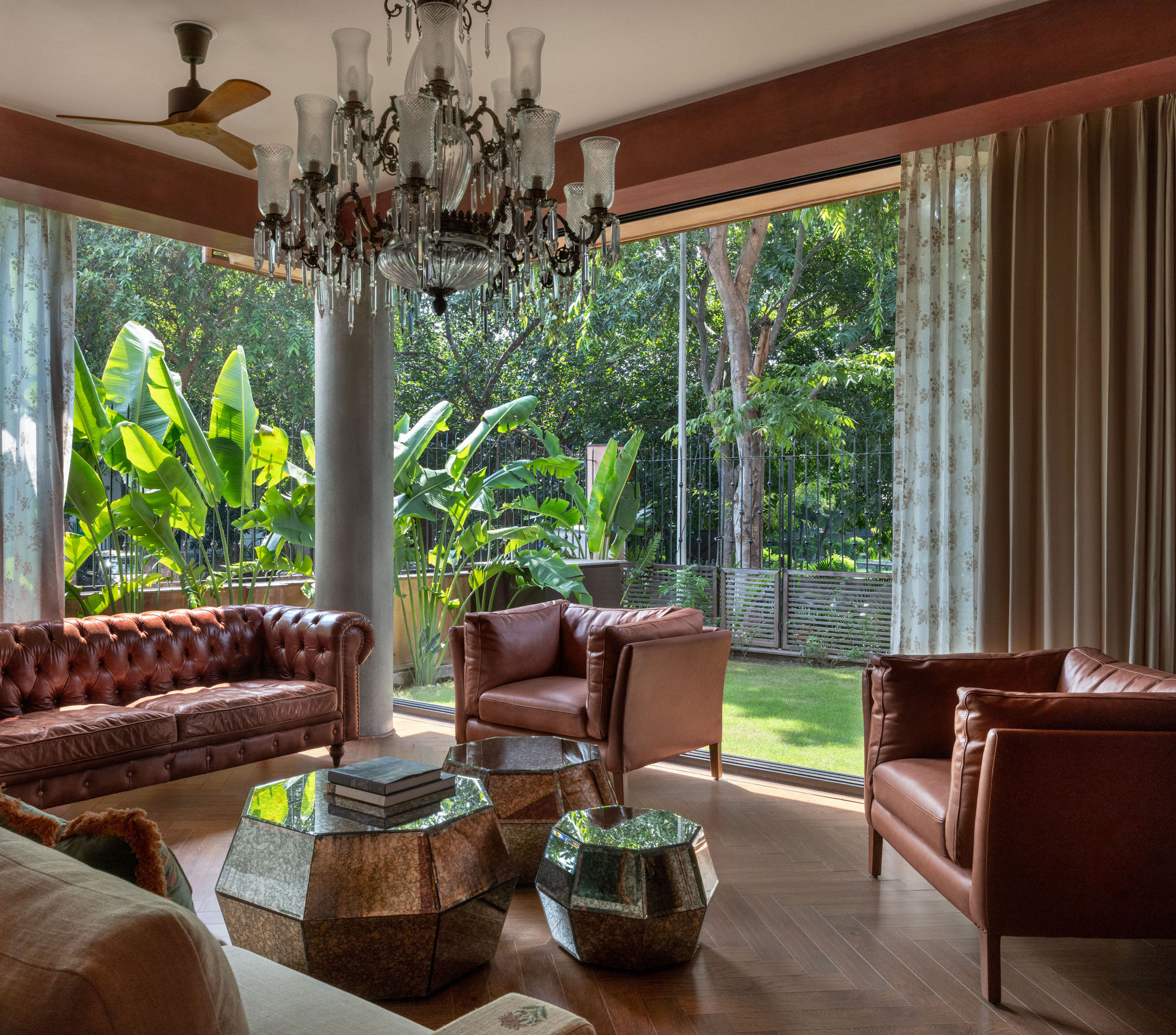 Living room with tan leather sofas, faceted mirrored tables and a chandelier, opening through floor-to-ceiling glass to banana plants and green lawn