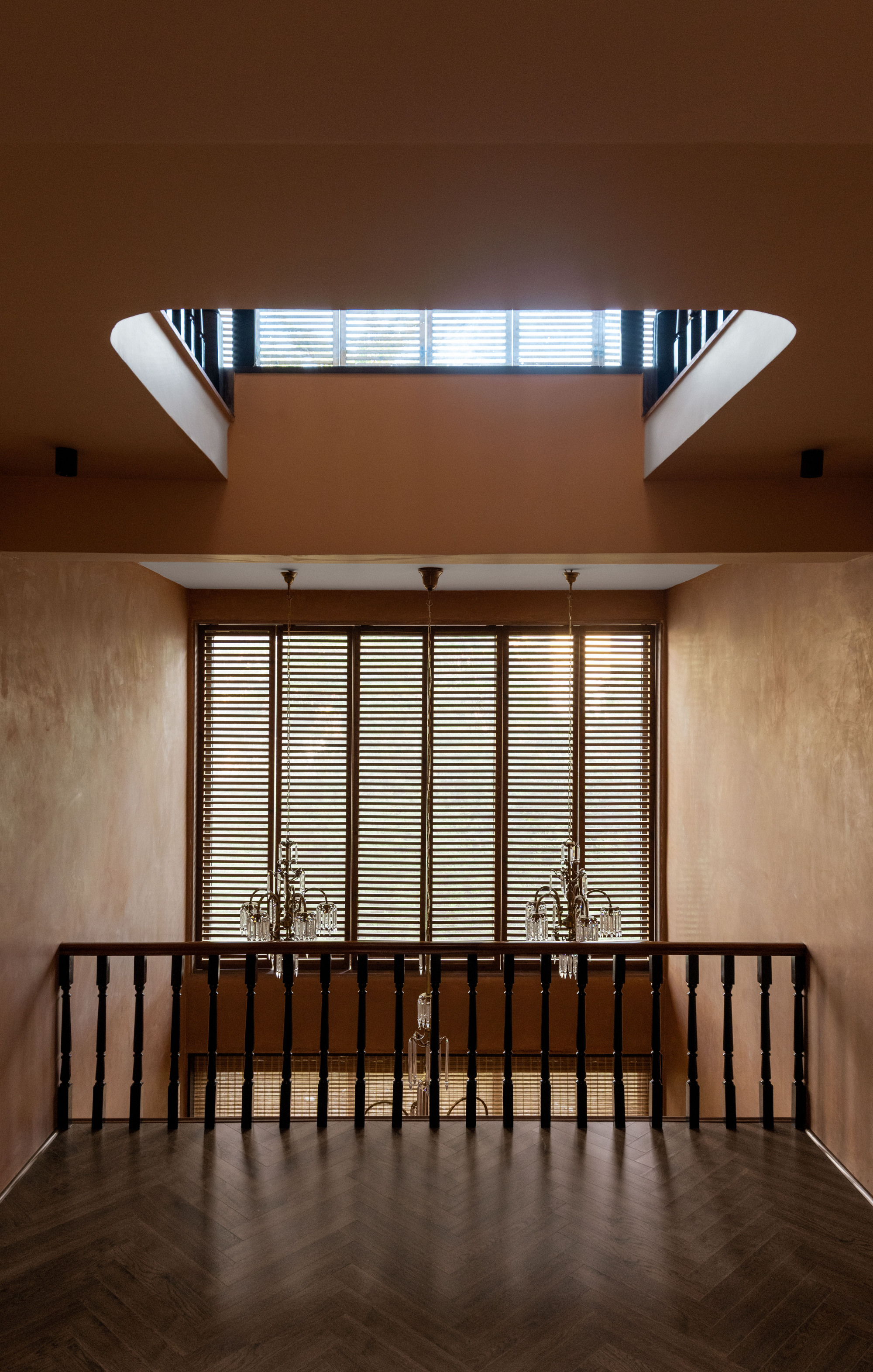 Stair landing with turned black balusters, slatted timber shutters and twin crystal chandeliers beneath a skylight, lit by daylight on herringbone flooring