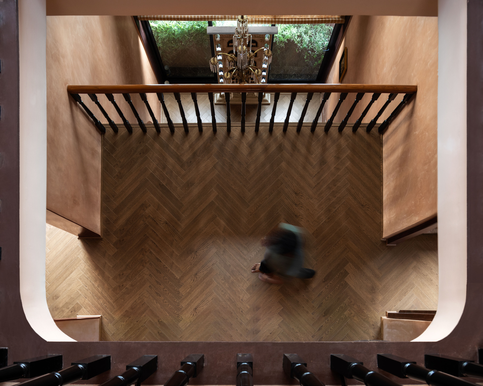 Top-down view of a herringbone timber floor with a blurred figure crossing below a baluster rail and chandelier