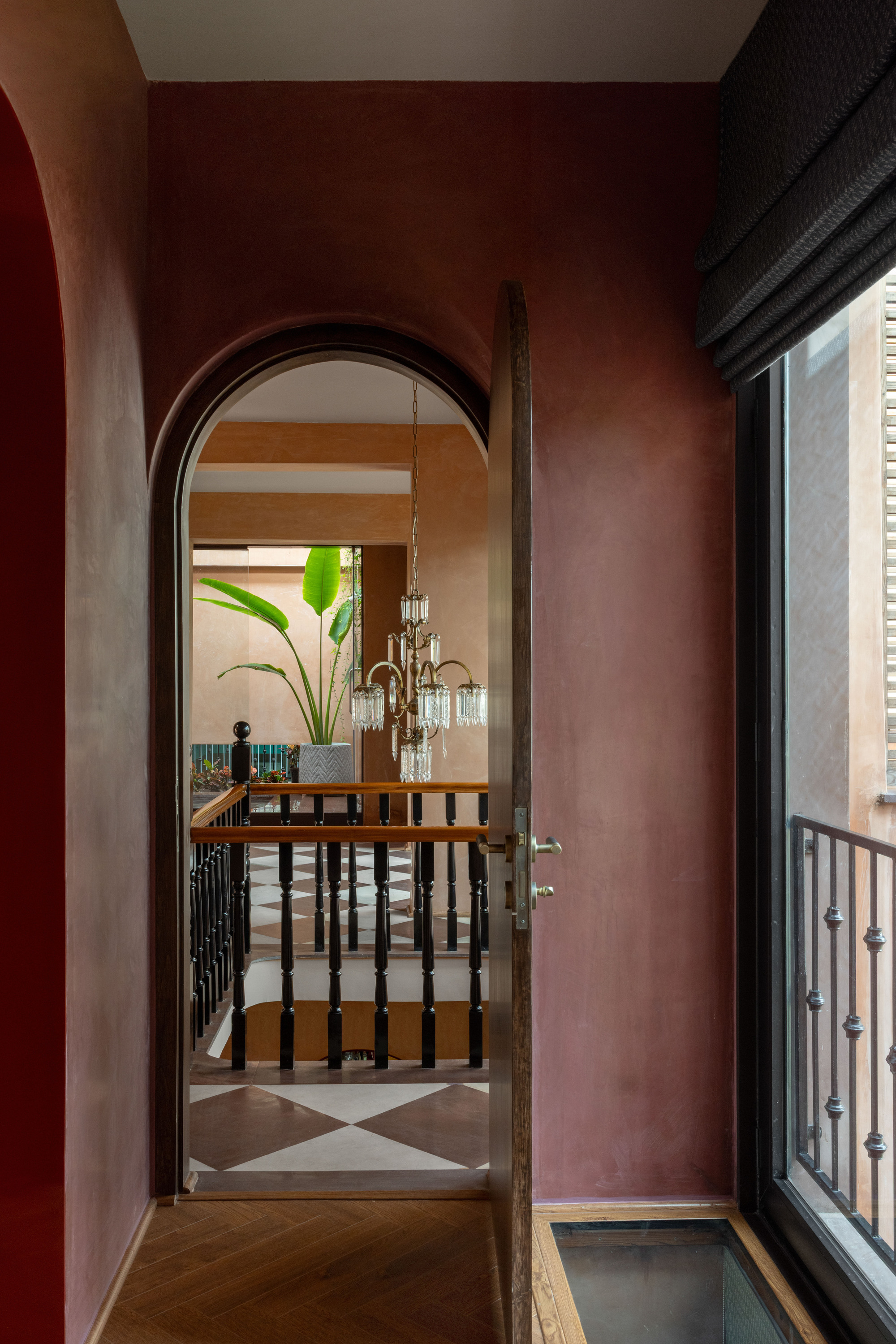 Arched timber door opening onto a landing with a chequered floor, banana plant and crystal chandelier, beside a window with a black blind