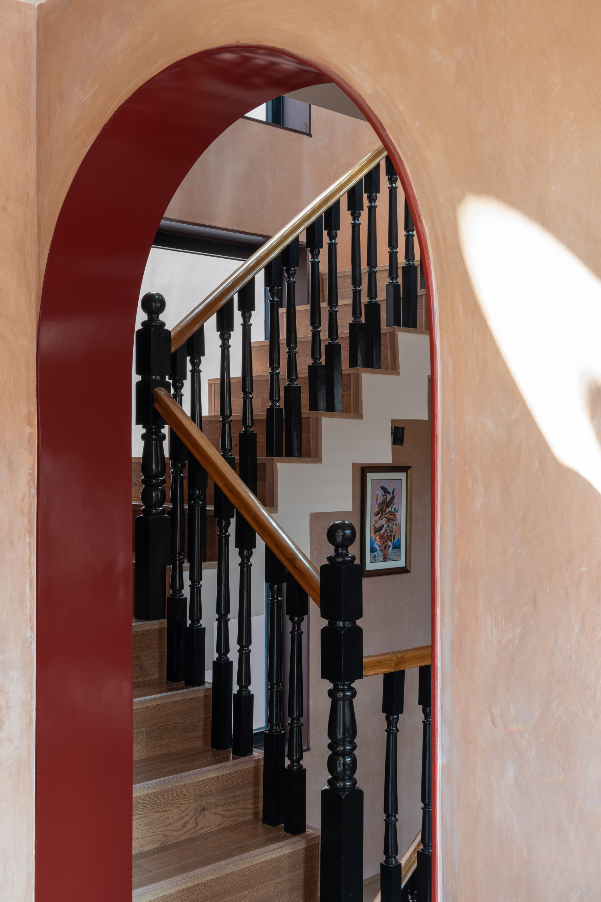 Red-edged arched opening framing a timber staircase with black turned balusters, a framed picture and a patch of afternoon sunlight on the wall