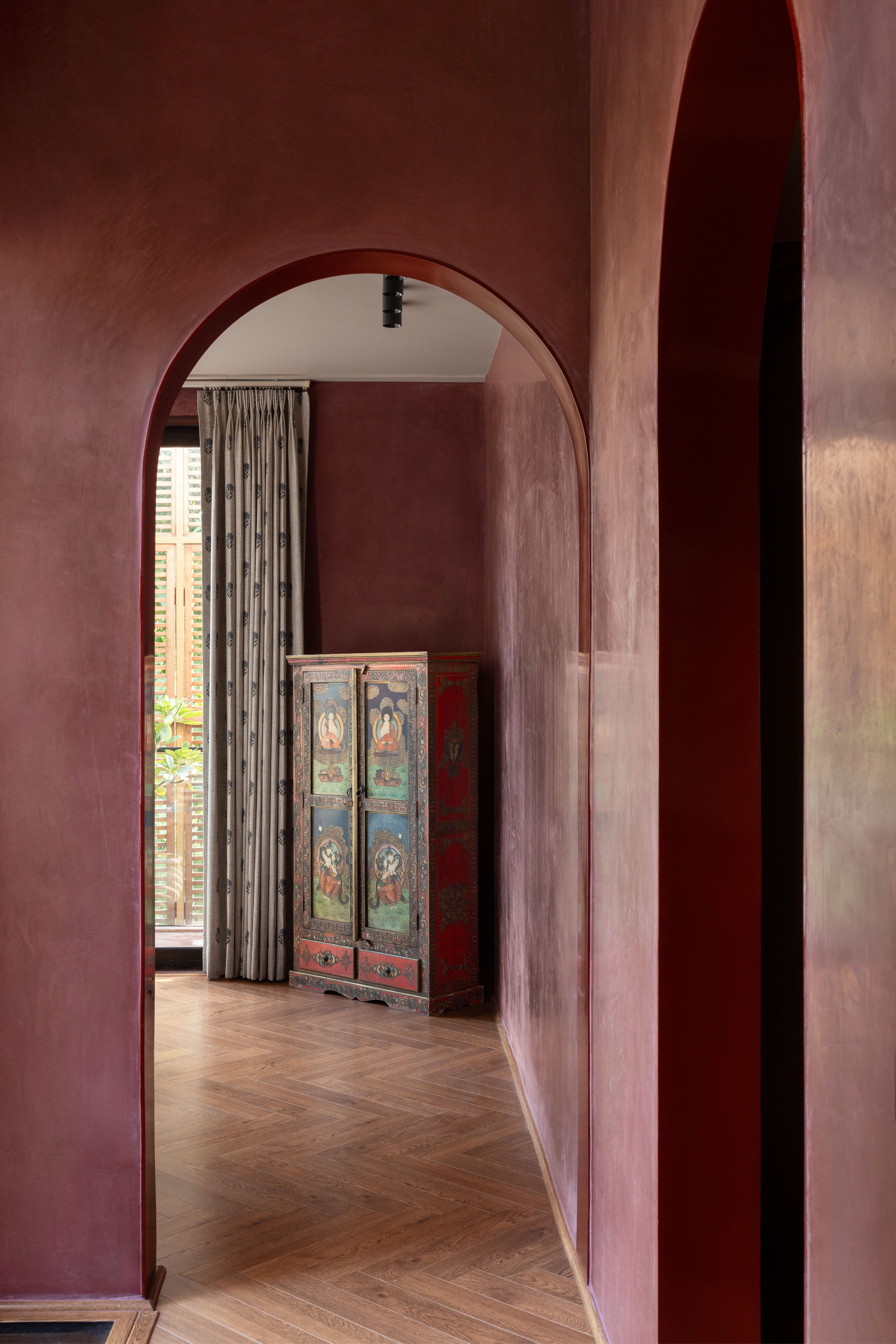 Arched passage in plum plaster walls leading to a painted wooden cabinet and a patterned curtain beside a shuttered window