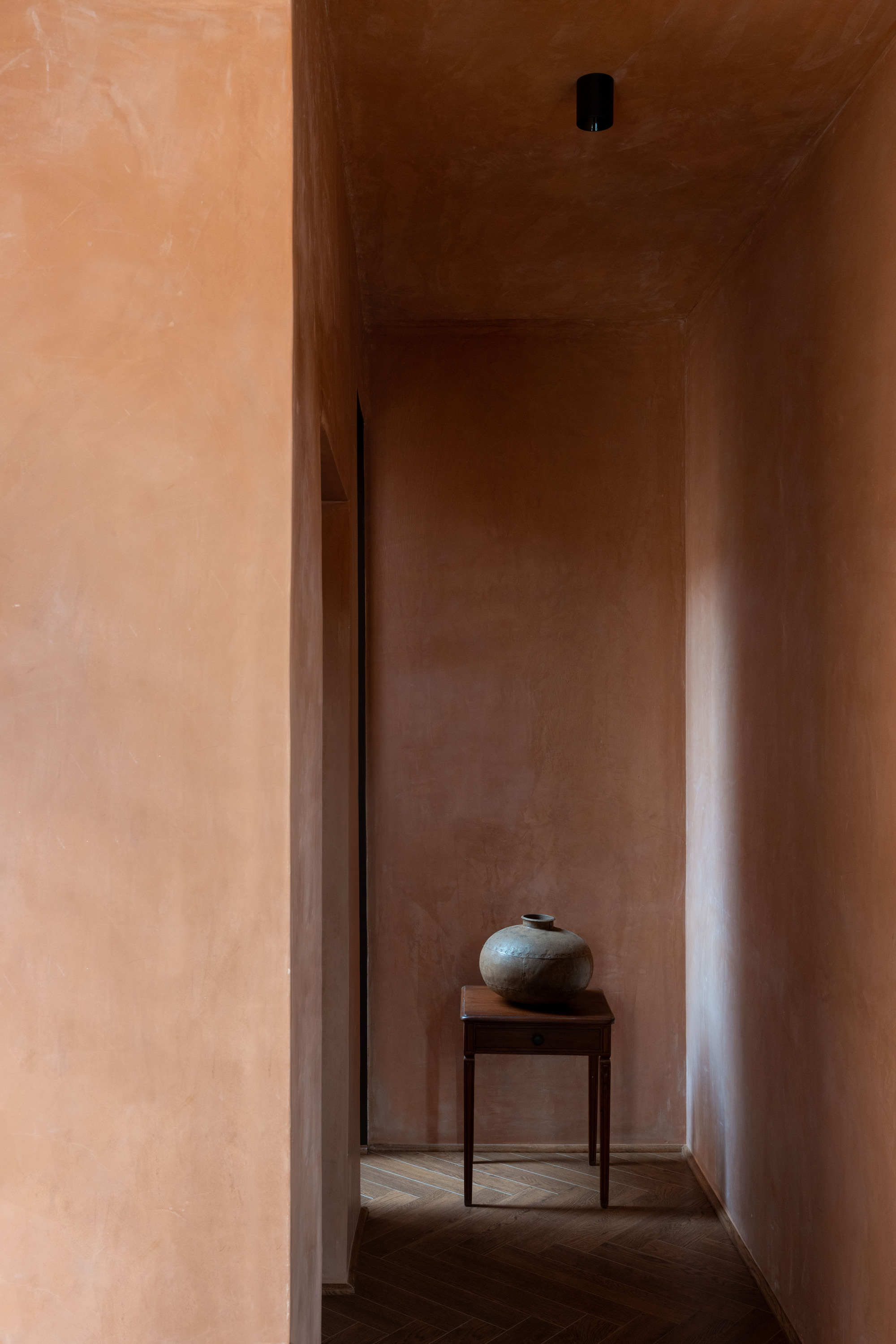 Narrow terracotta-plastered corridor ending in a small wooden side table holding a clay pot, with a black ceiling spotlight above