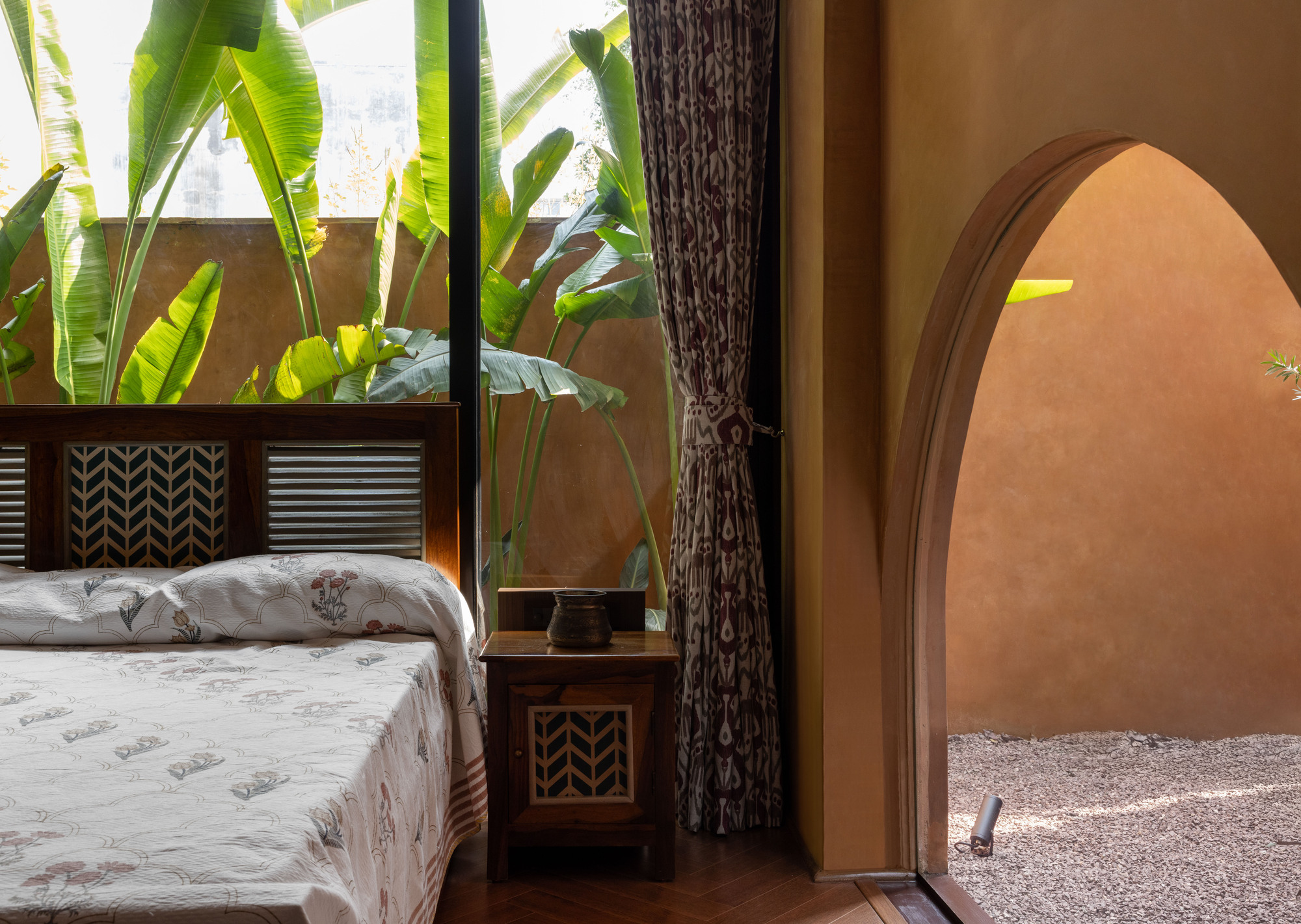 Bedroom corner with a carved timber bed and bedside table beside an arched opening, banana leaves pressing against the glass