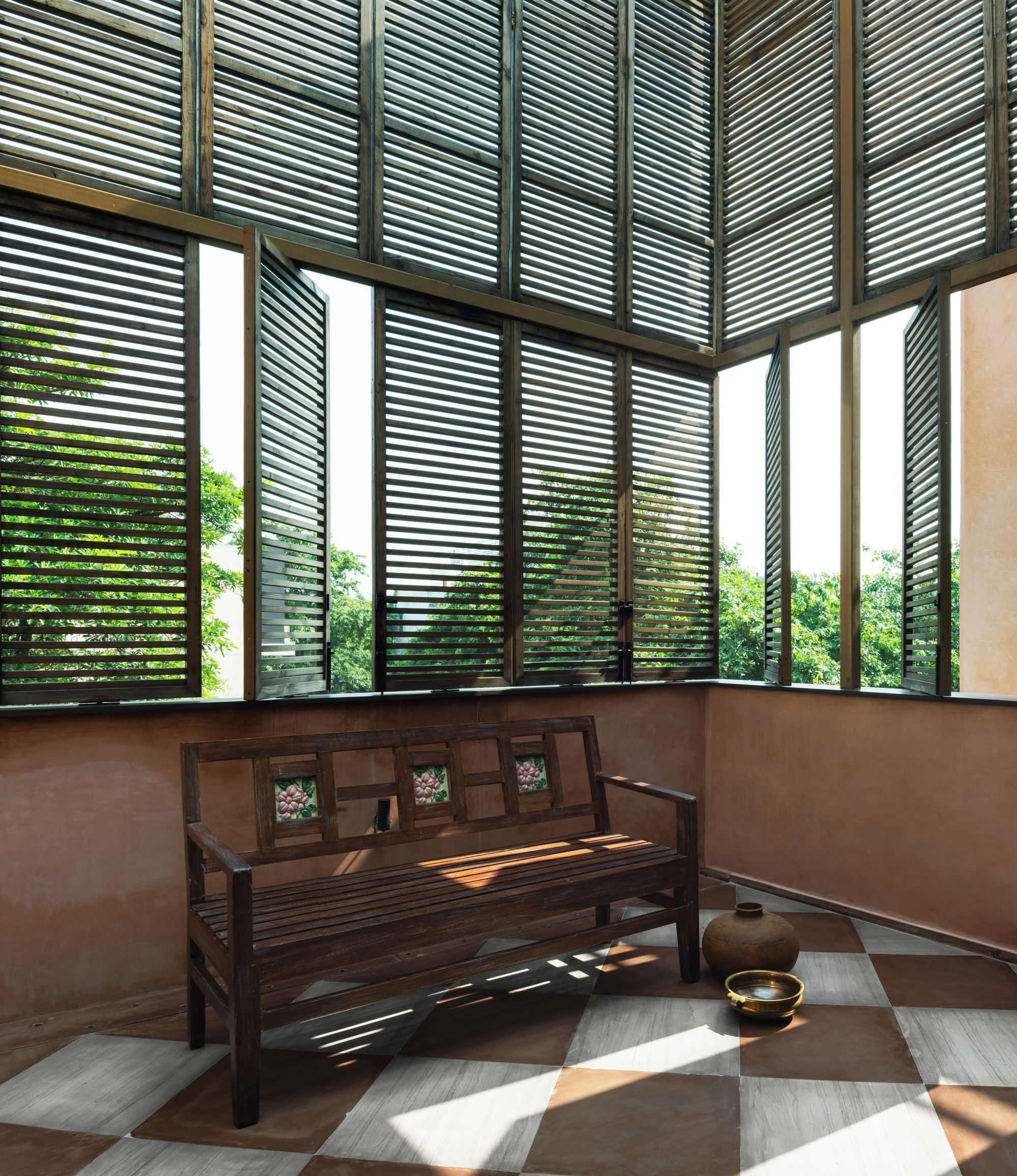 Wooden bench with inlaid tile backrest in a corner of louvered shutters, sunlight striping a checkered stone floor
