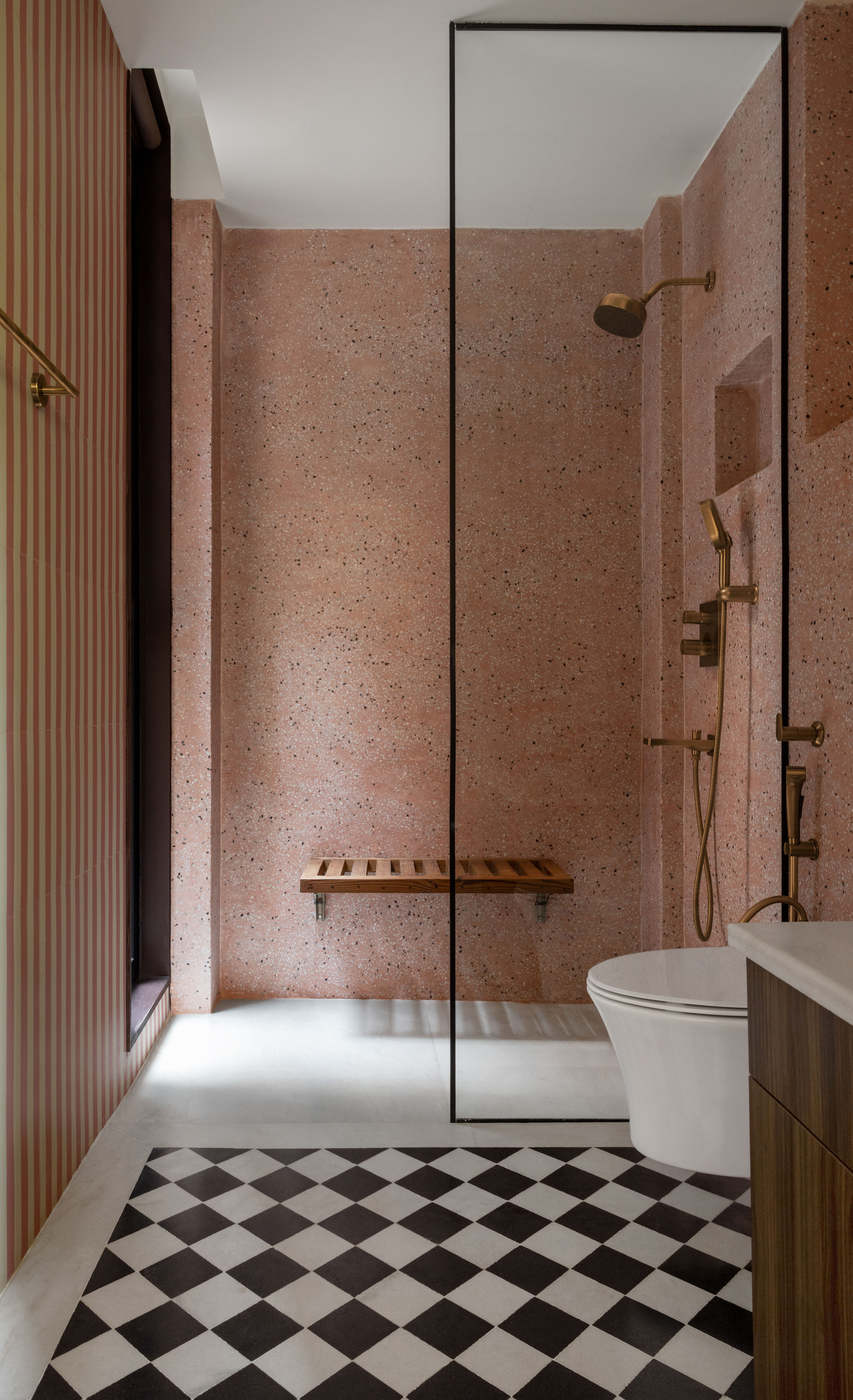 Bathroom with pink terrazzo shower wall, teak fold-down bench and brass fittings above a black and white checkerboard floor