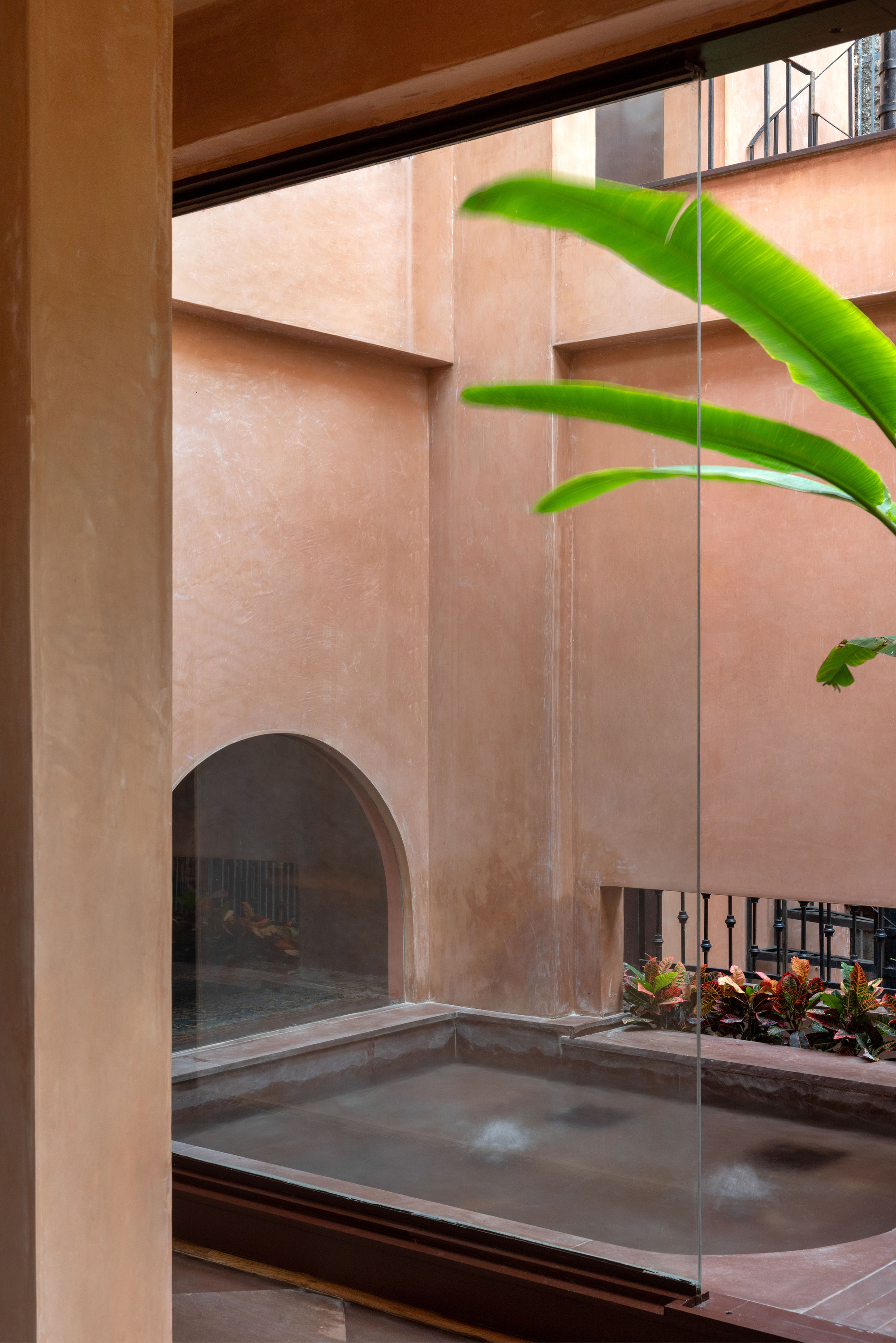 View through a glass pane to a small plunge pool in a terracotta plaster courtyard with a banana leaf overhead