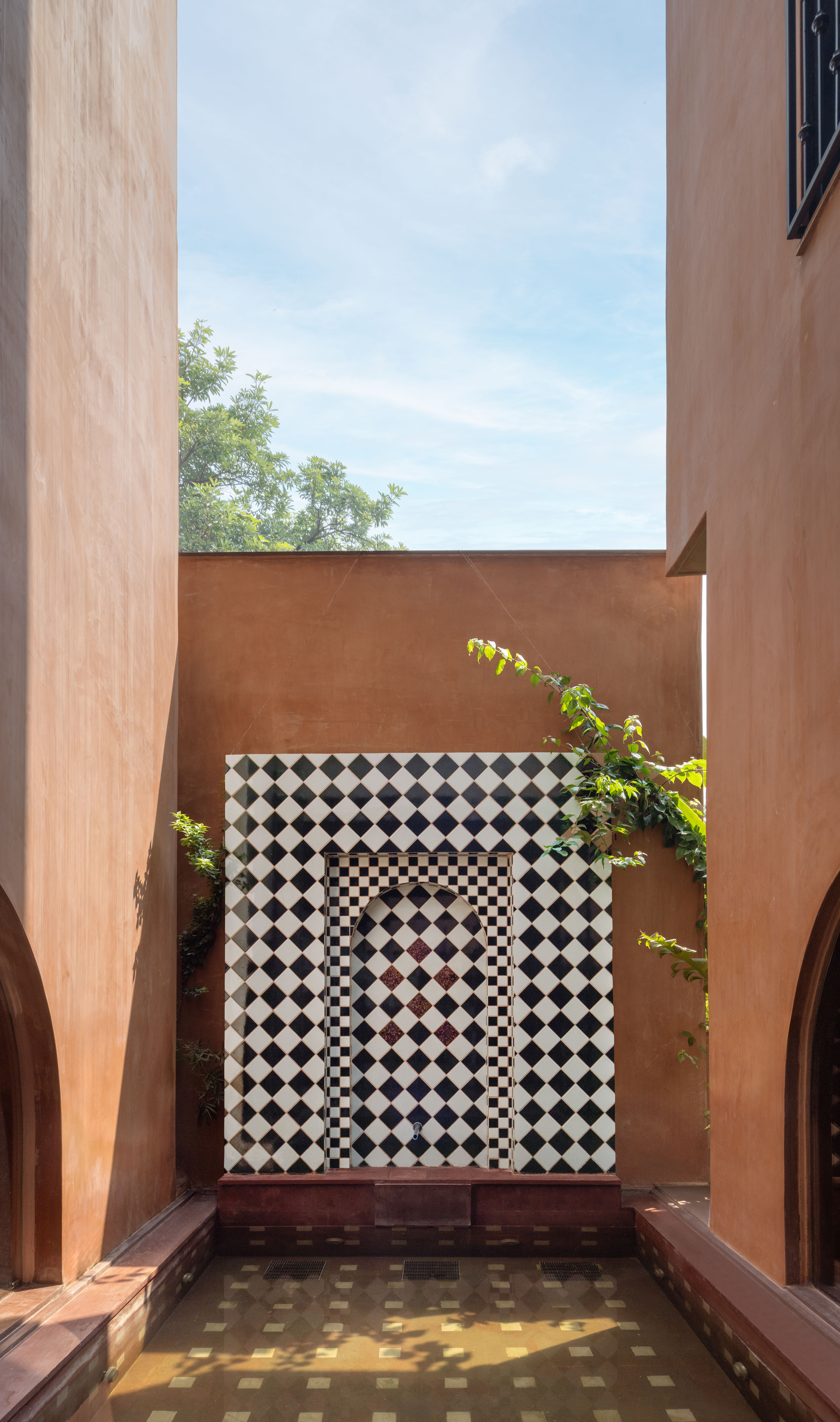 Courtyard fountain niche clad in black and white diamond tile between tall ochre walls, with a tree against a blue sky