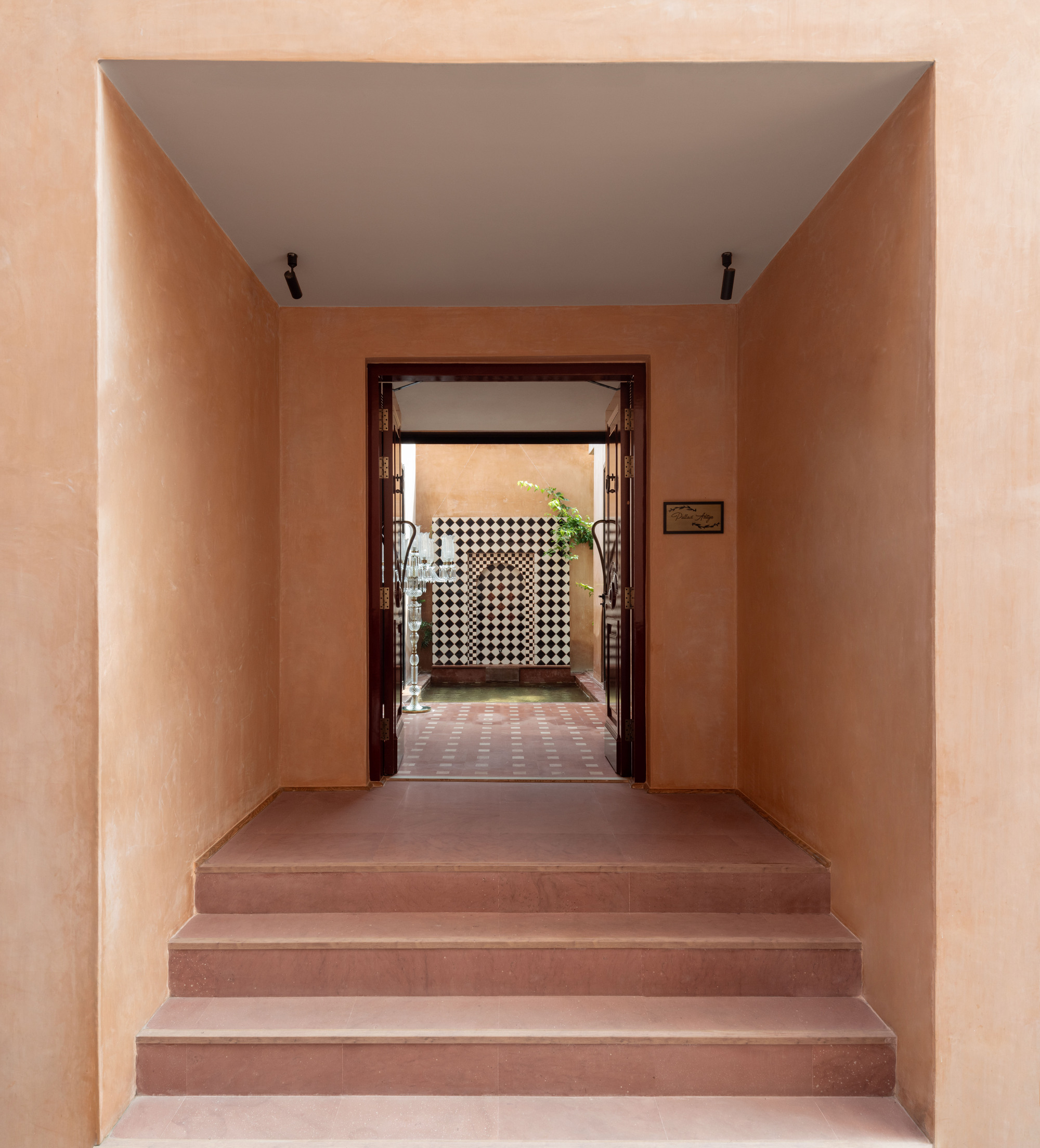 Red sandstone steps rising to a recessed terracotta-plastered entrance, with a checkered tile wall and green plant glimpsed through open timber doors