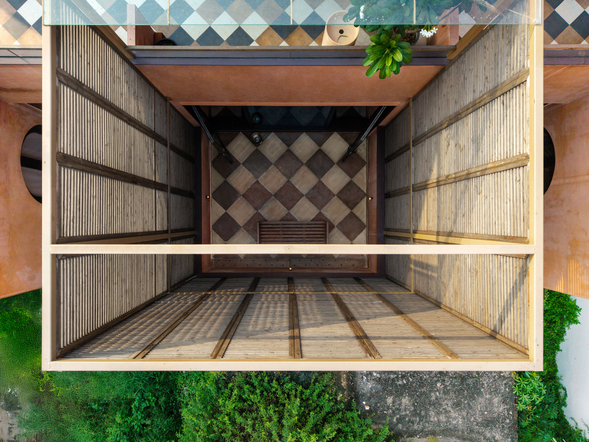 Top-down view of a slatted timber-clad void with a checkered tile floor and a potted plant at its edge, ringed by greenery