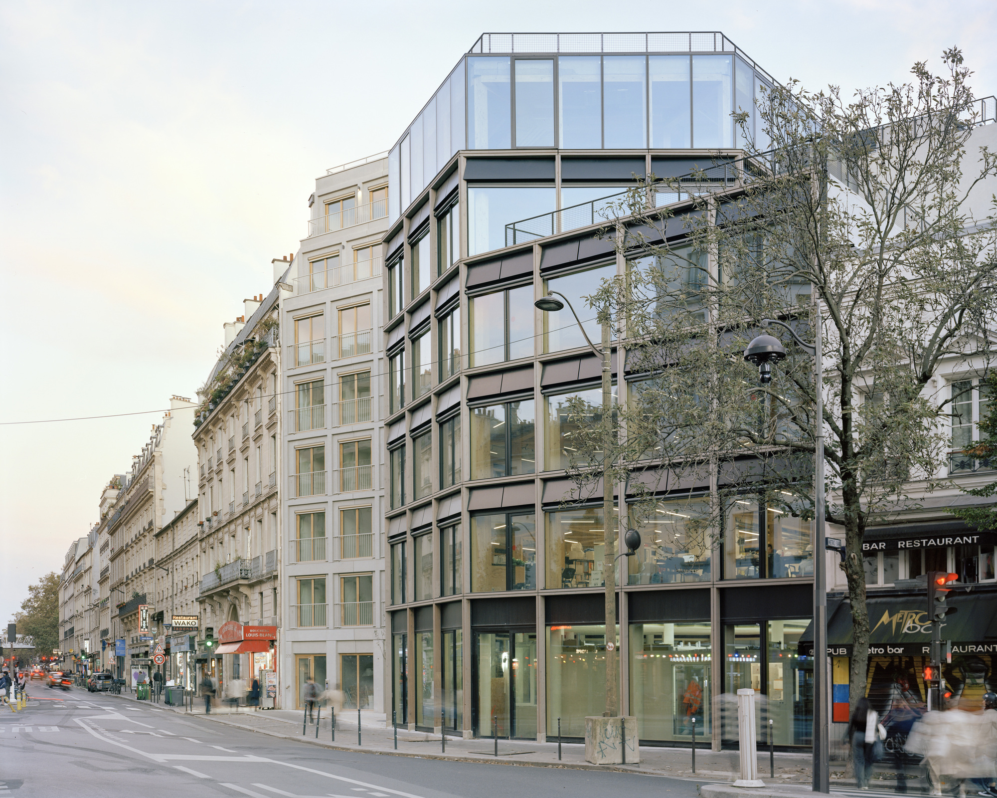 Corner of a glass and dark metal framed building beside stone apartment blocks, with a bare plane tree and blurred pedestrians at dusk