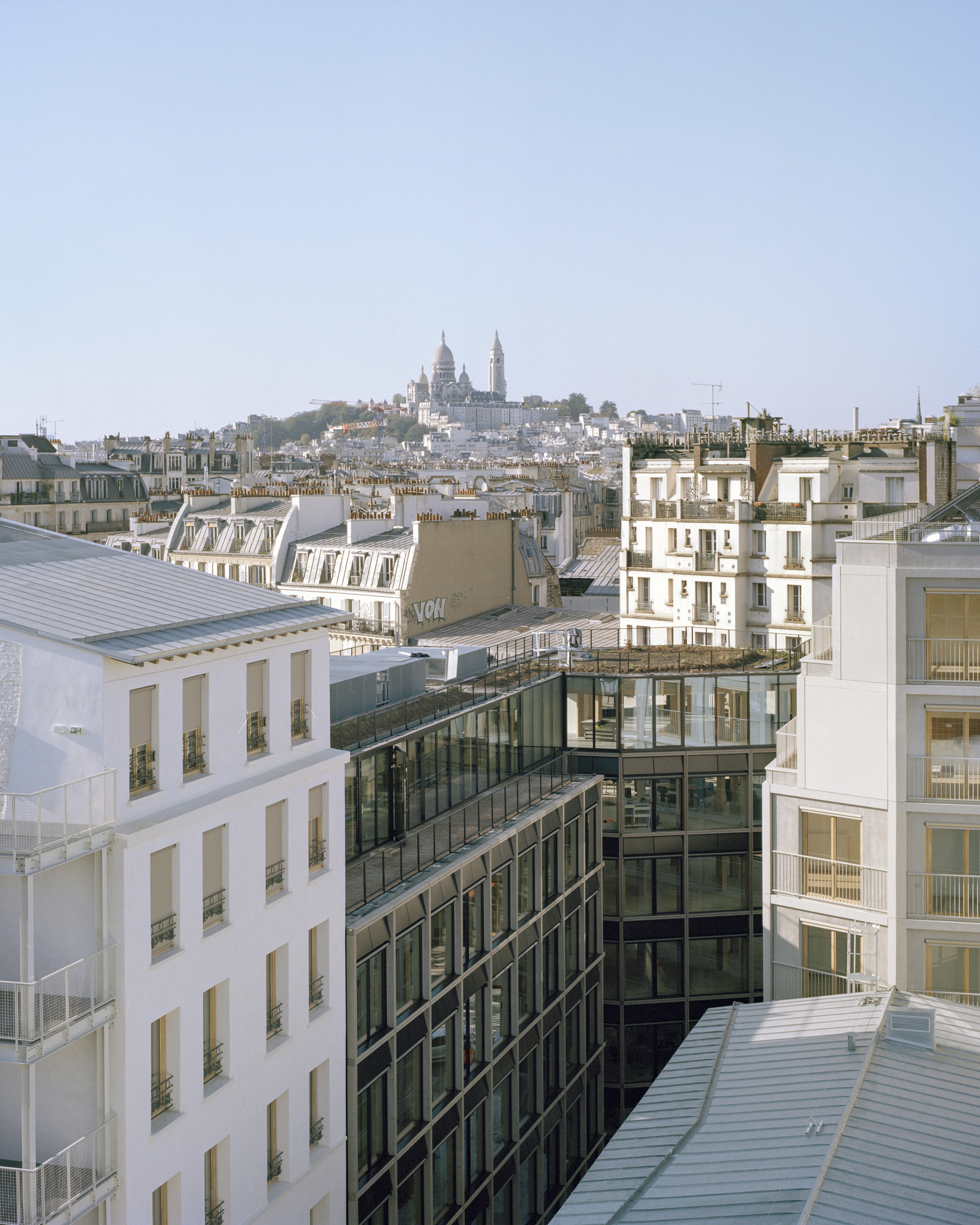 Rooftop view over zinc roofs and a dark glazed facade toward a distant white domed church on a hill in clear haze