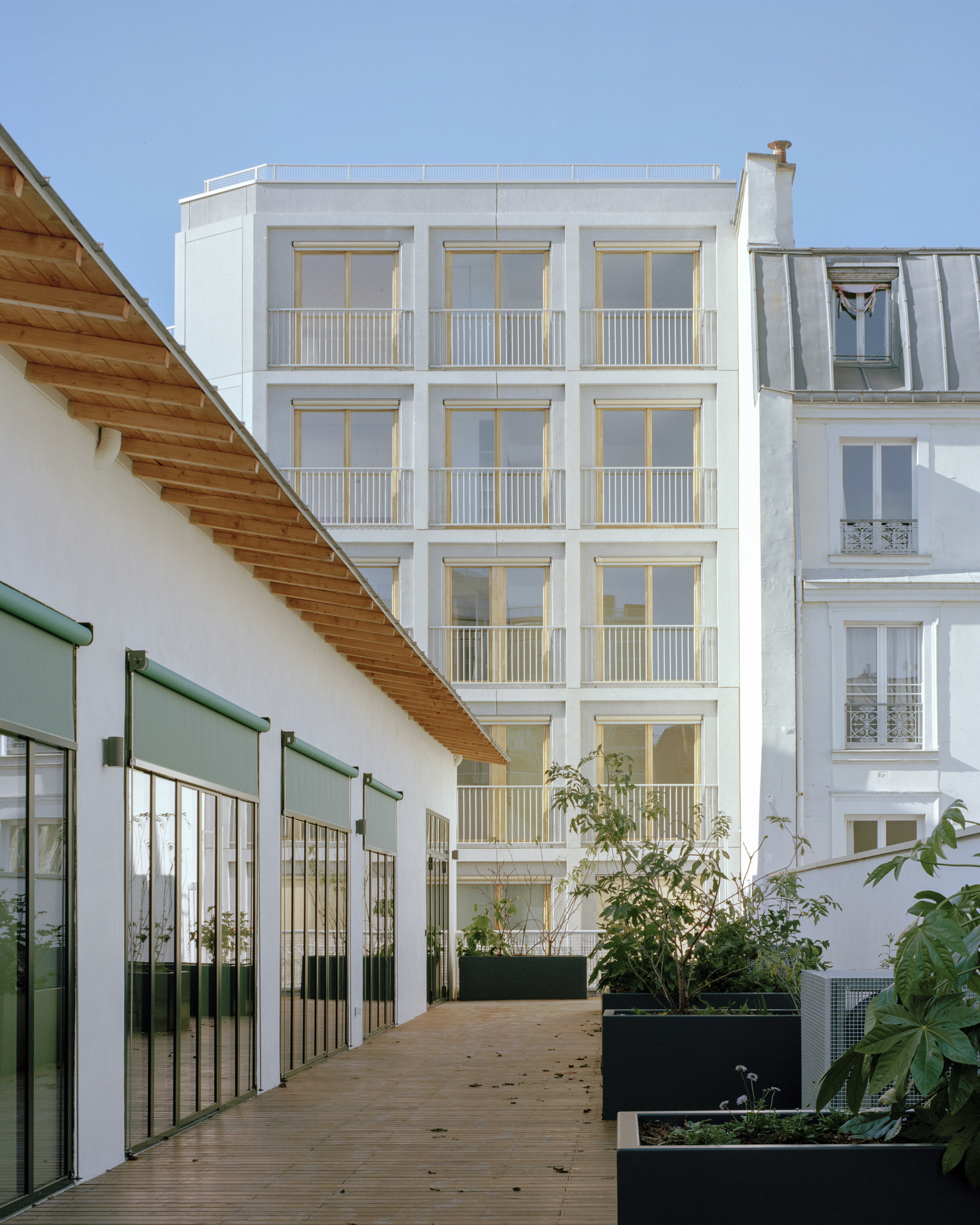 Timber-roofed terrace with a wooden deck and black planters, facing a white balconied apartment block under a clear blue sky