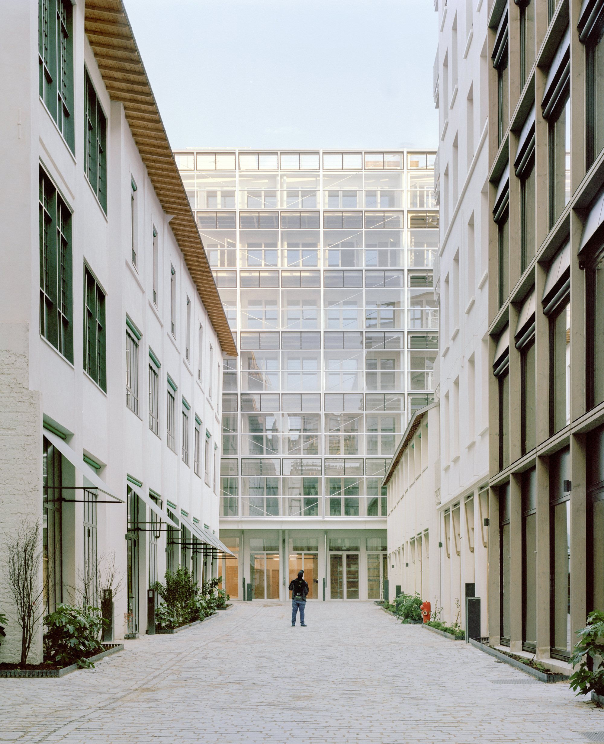 Cobbled passage between white and timber-framed facades leading to a glazed block, with a lone figure standing in the middle of the paving